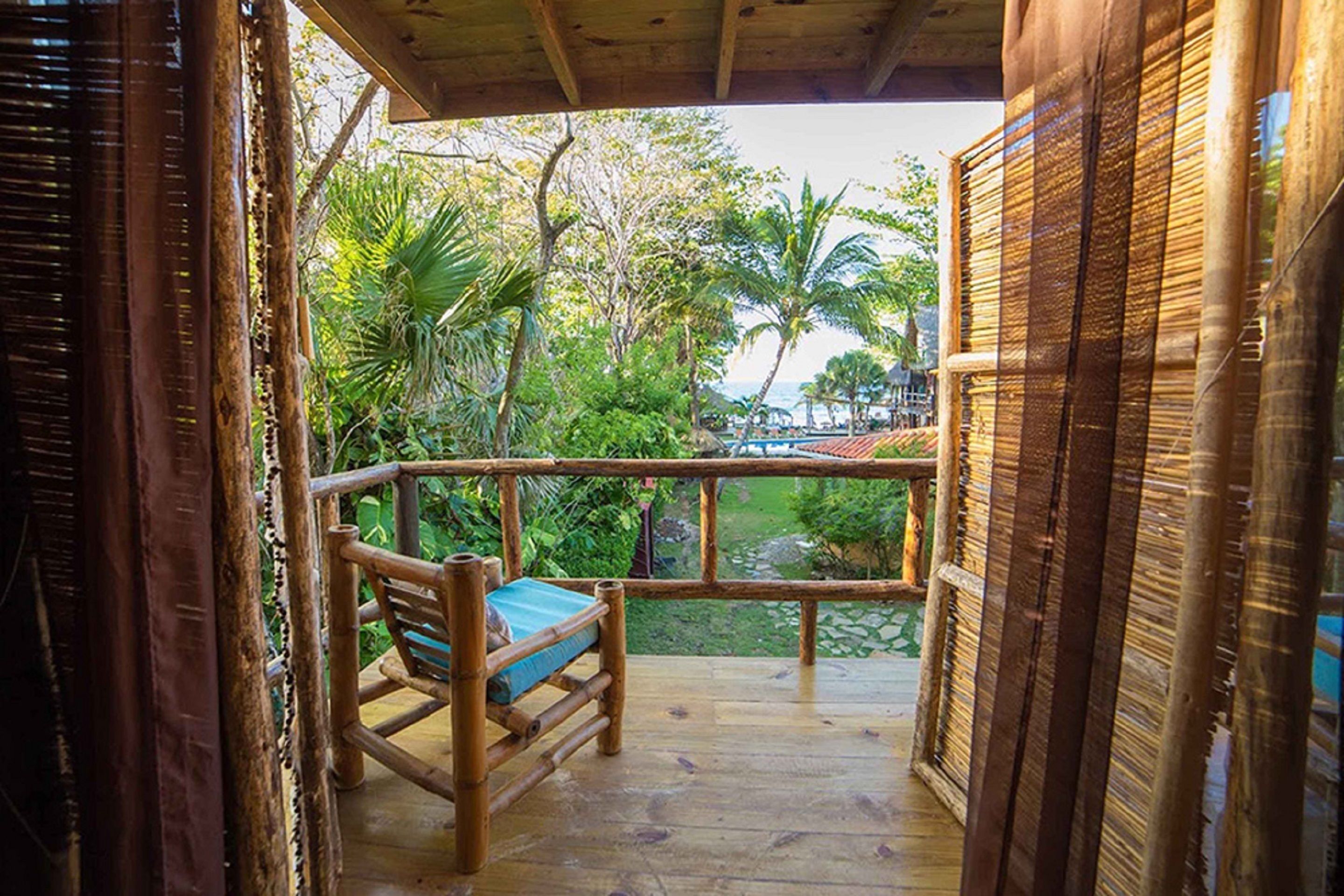 A wooden deck with a bamboo railing, a blue cushioned bench, and lush greenery in the background.