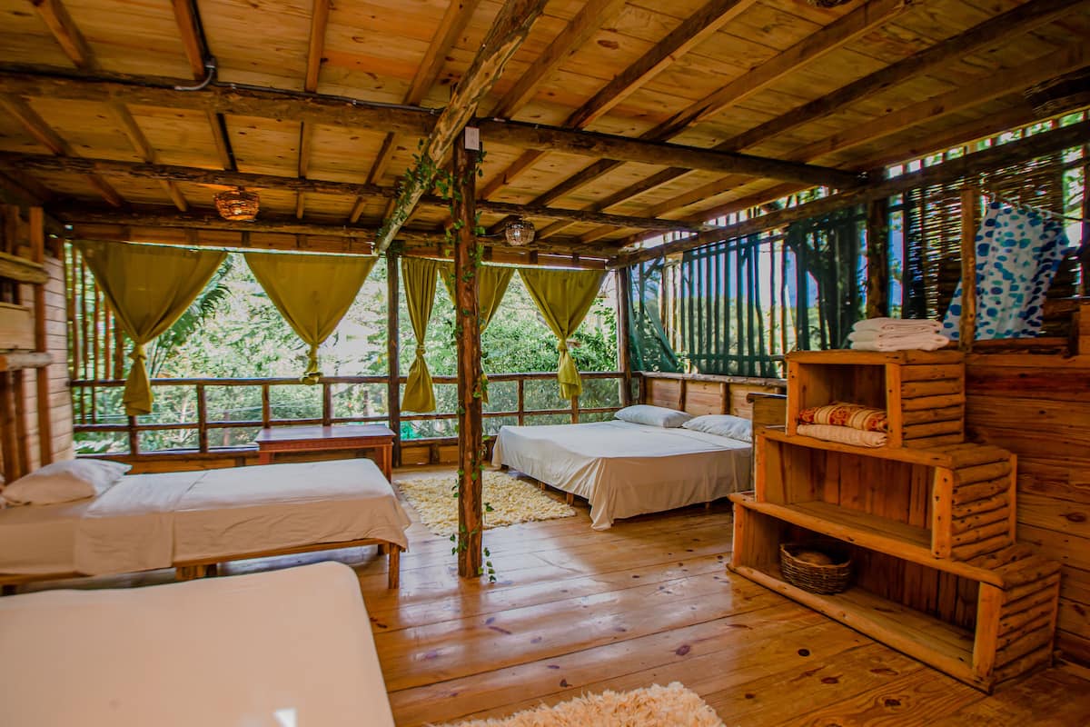 Una cabaña de madera con dos camas, un estante de madera con toallas y vistas a la vegetación a través de las ventanas.