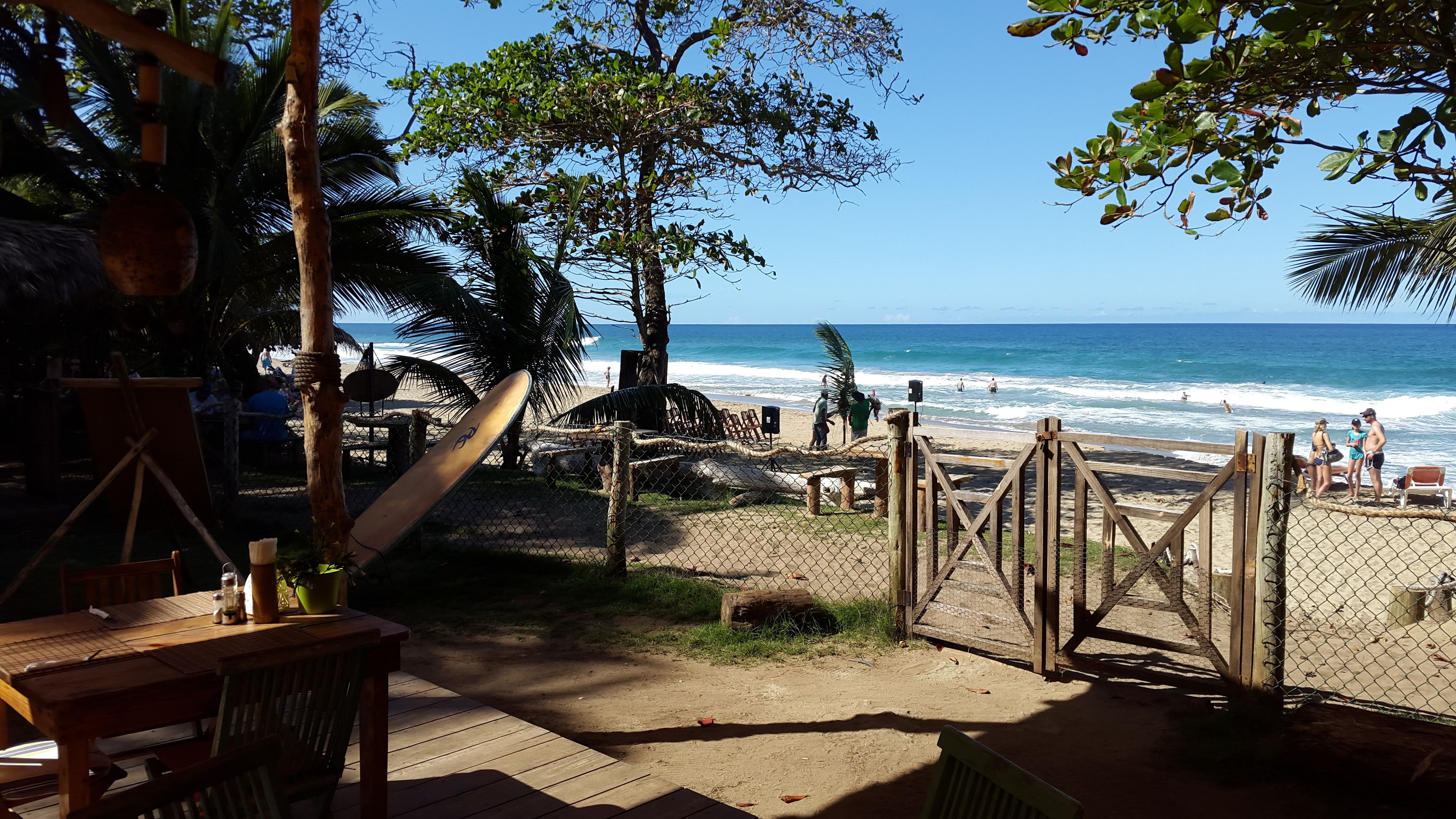 Vista de la playa con una terraza de madera, una tabla de surf y una valla que da al océano.