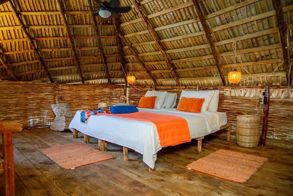 A rustic room with a thatched roof, a bed with orange and white bedding, and a woven basket on the floor.