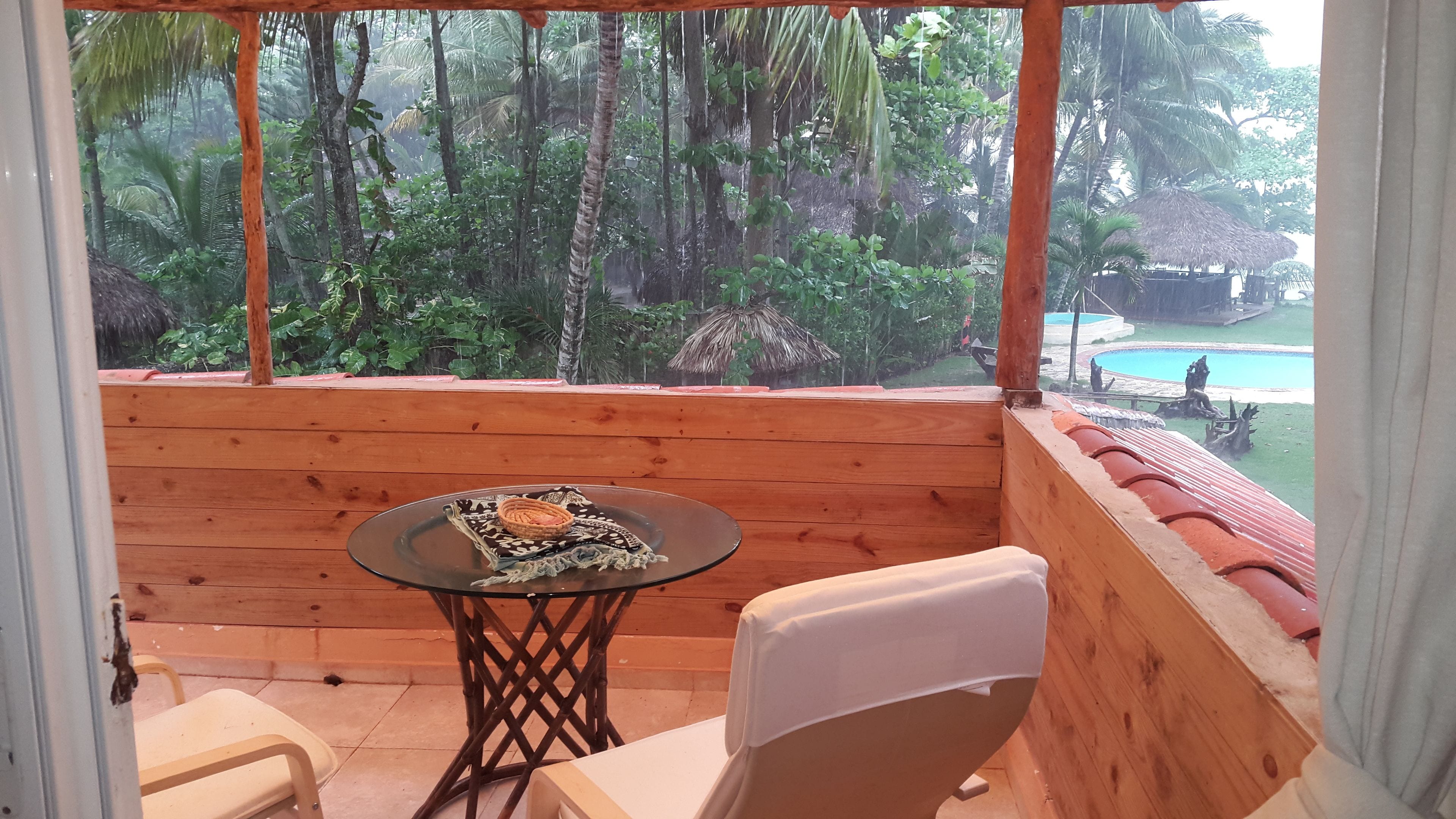 Un balcon en bois avec une table en verre, une chaise blanche et une vue sur une piscine et une végétation tropicale.