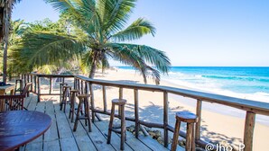 A wooden deck with tables and stools overlooking a beach with palm trees and the ocean.