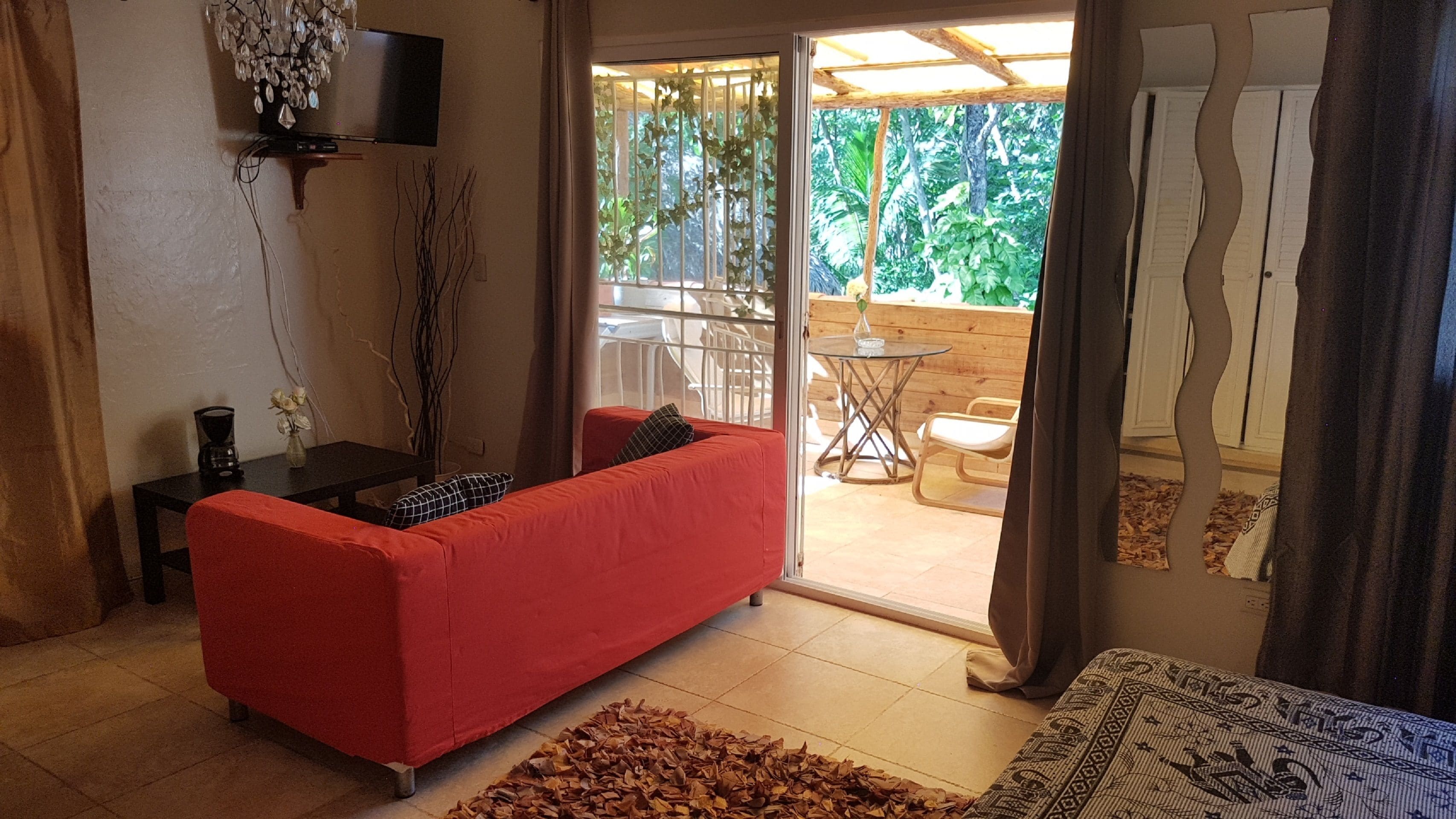A living room with a red sofa, a TV mounted on the wall, and a view of an outdoor patio area with a table and chairs.