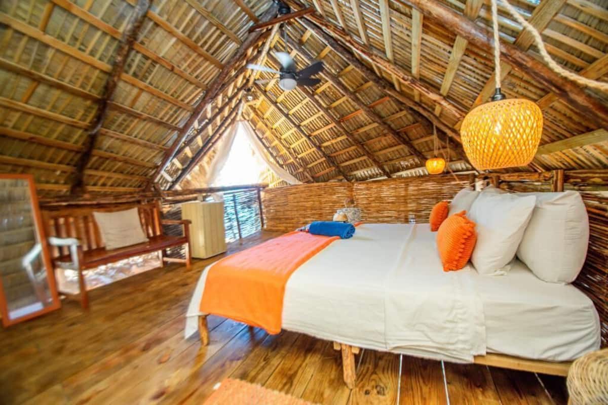Una habitación con techo de madera alto, cama con ropa de cama blanca y naranja, y una lámpara colgante de fibra natural.