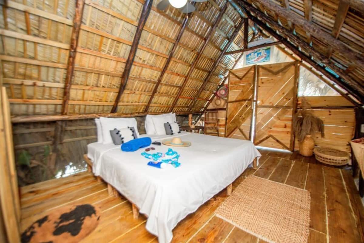 Una habitación rústica y acogedora con techo y paredes de madera, una cama con ropa blanca y un tapete tejido en el suelo.
