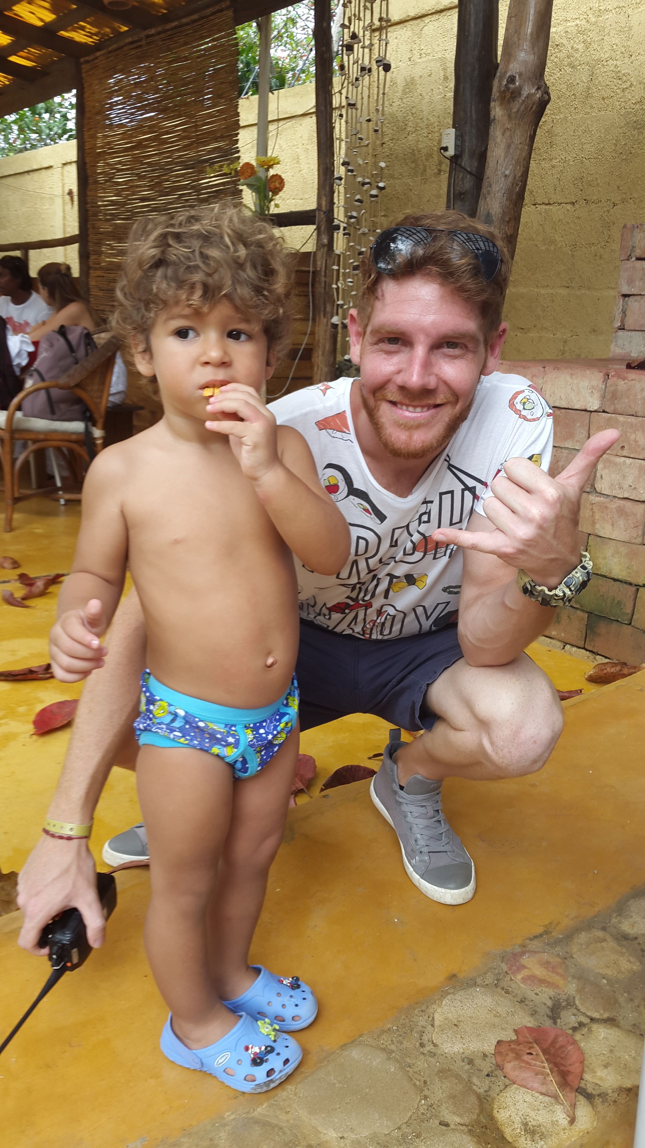 A man and a child in a casual outdoor setting, possibly a patio or courtyard, with a yellow floor and brick walls.