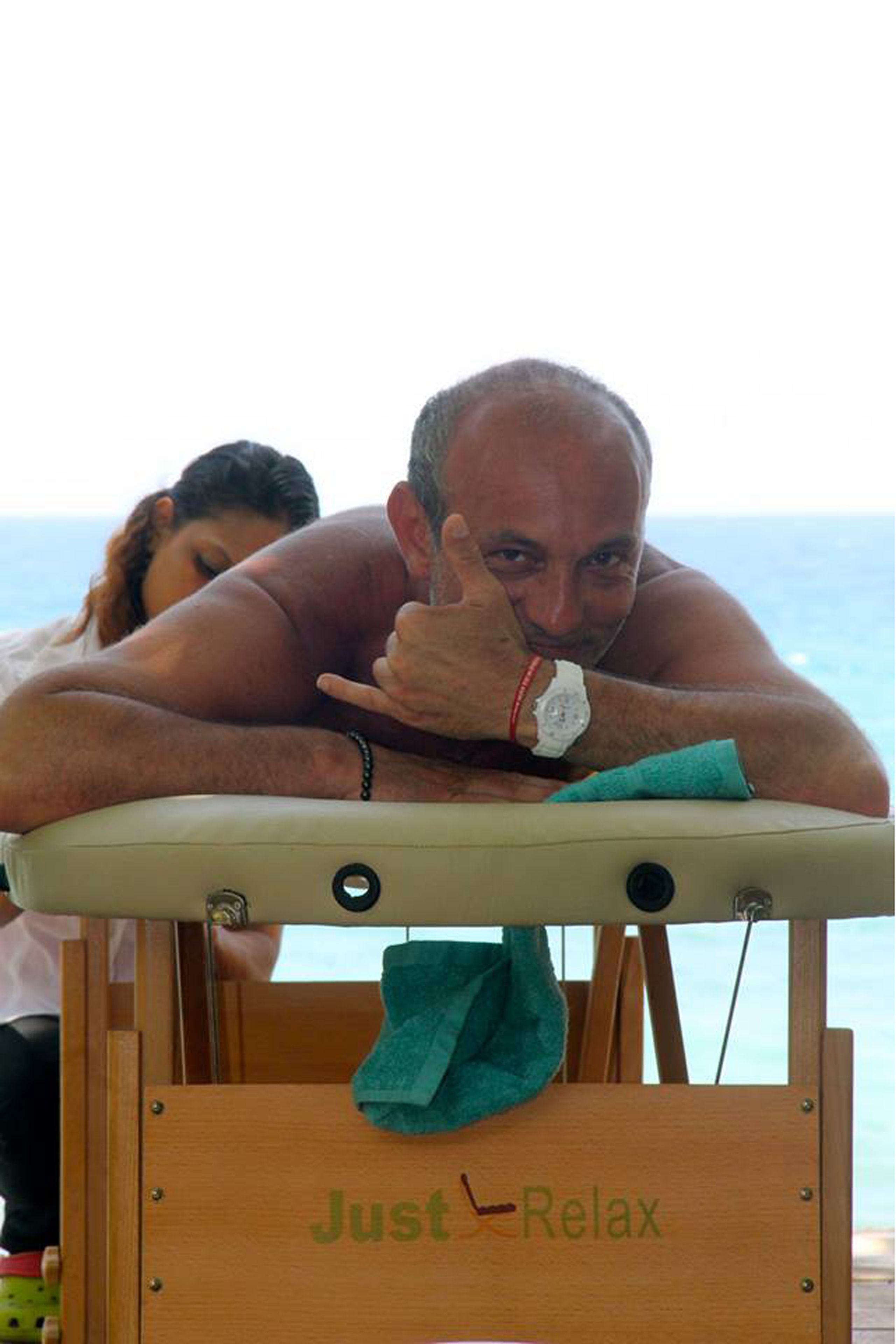 Un hombre recuesta la cabeza sobre una silla de madera con una toalla, mirando un cuerpo de agua.