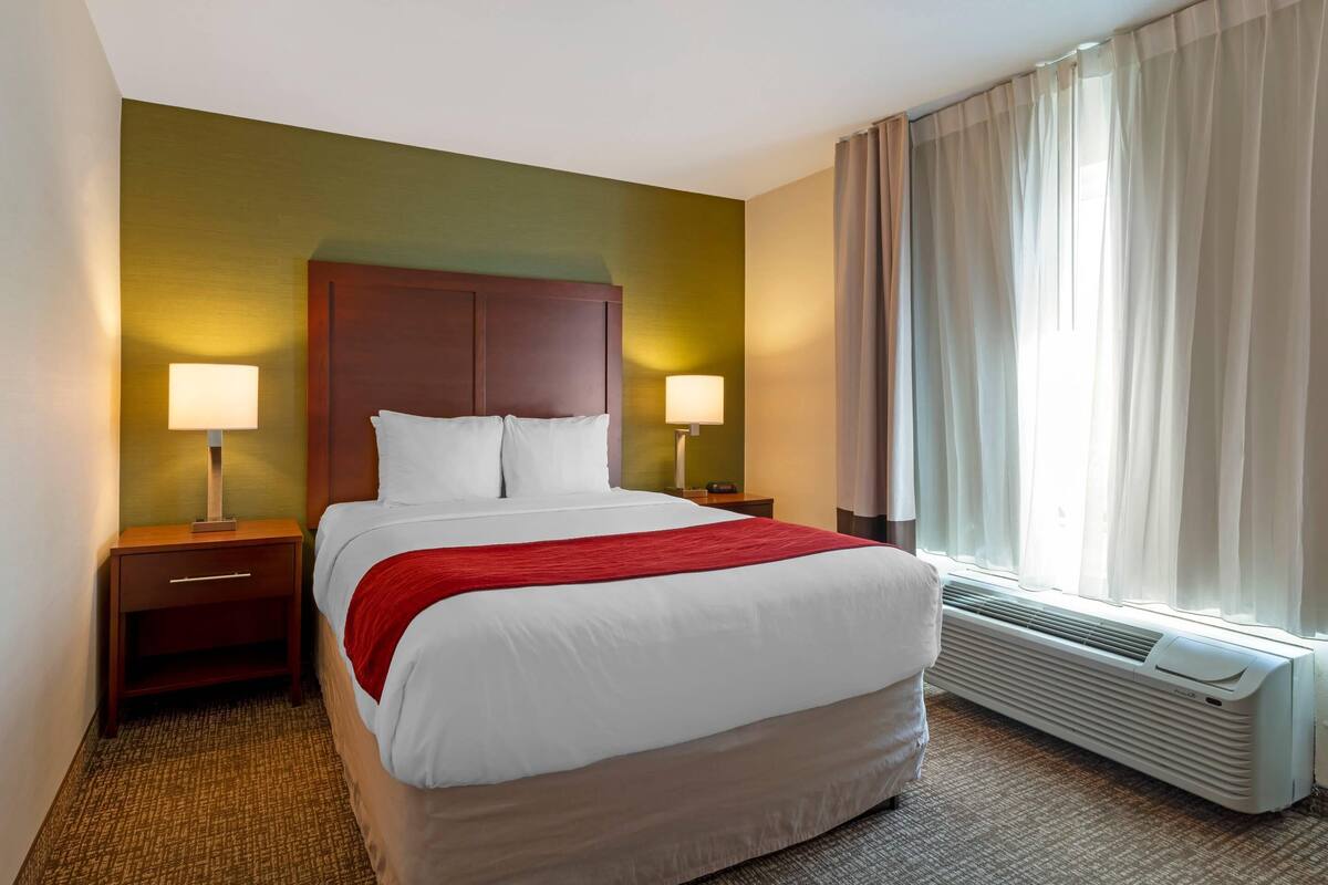 Una habitación de hotel con una cama, dos mesitas de noche con lámparas, una ventana con cortinas y un aire acondicionado instalado en la pared.