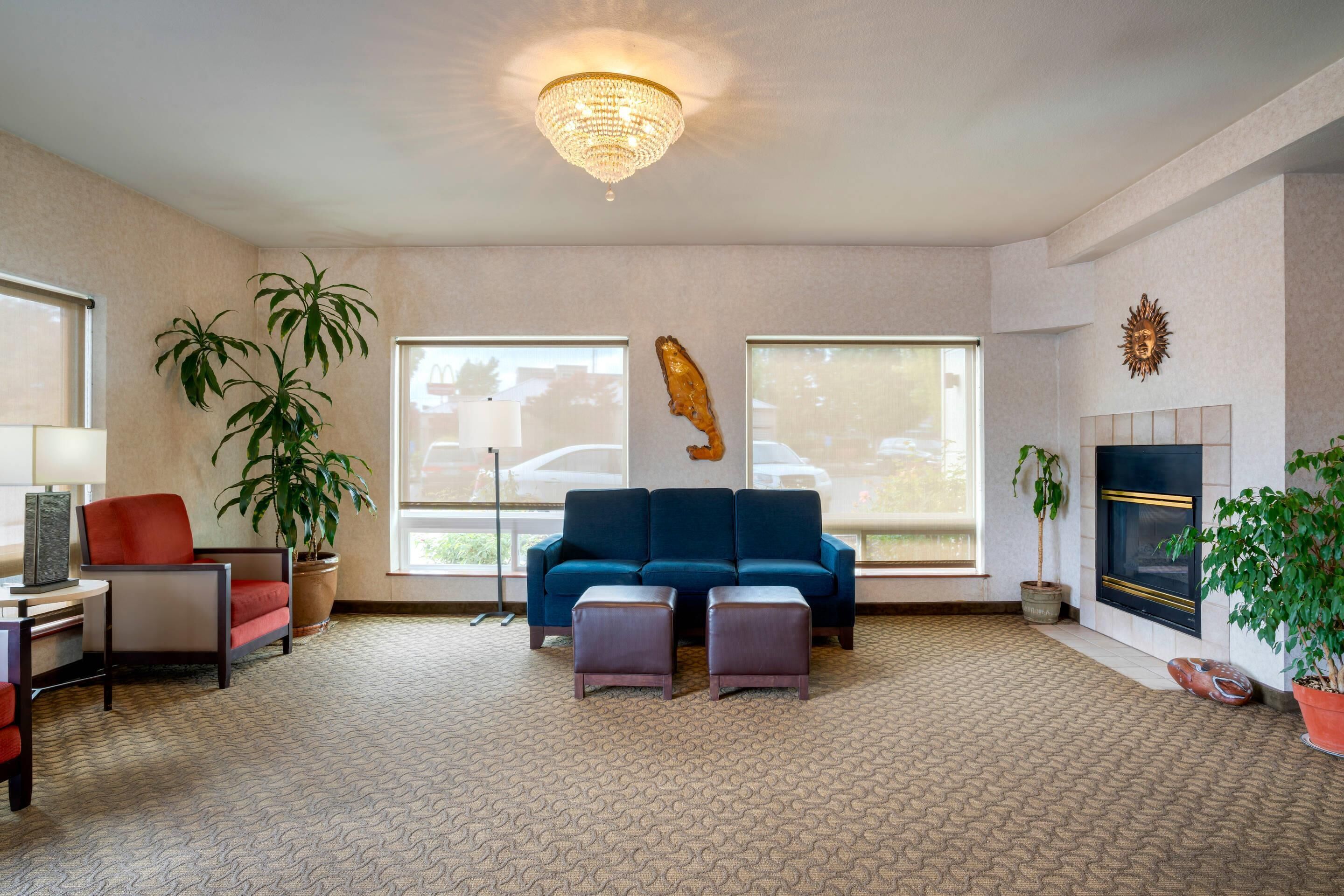 A modern living room with a blue sofa, two ottomans, a red chair, a desk, a lamp, a large potted plant, a fireplace, and a chandelier.