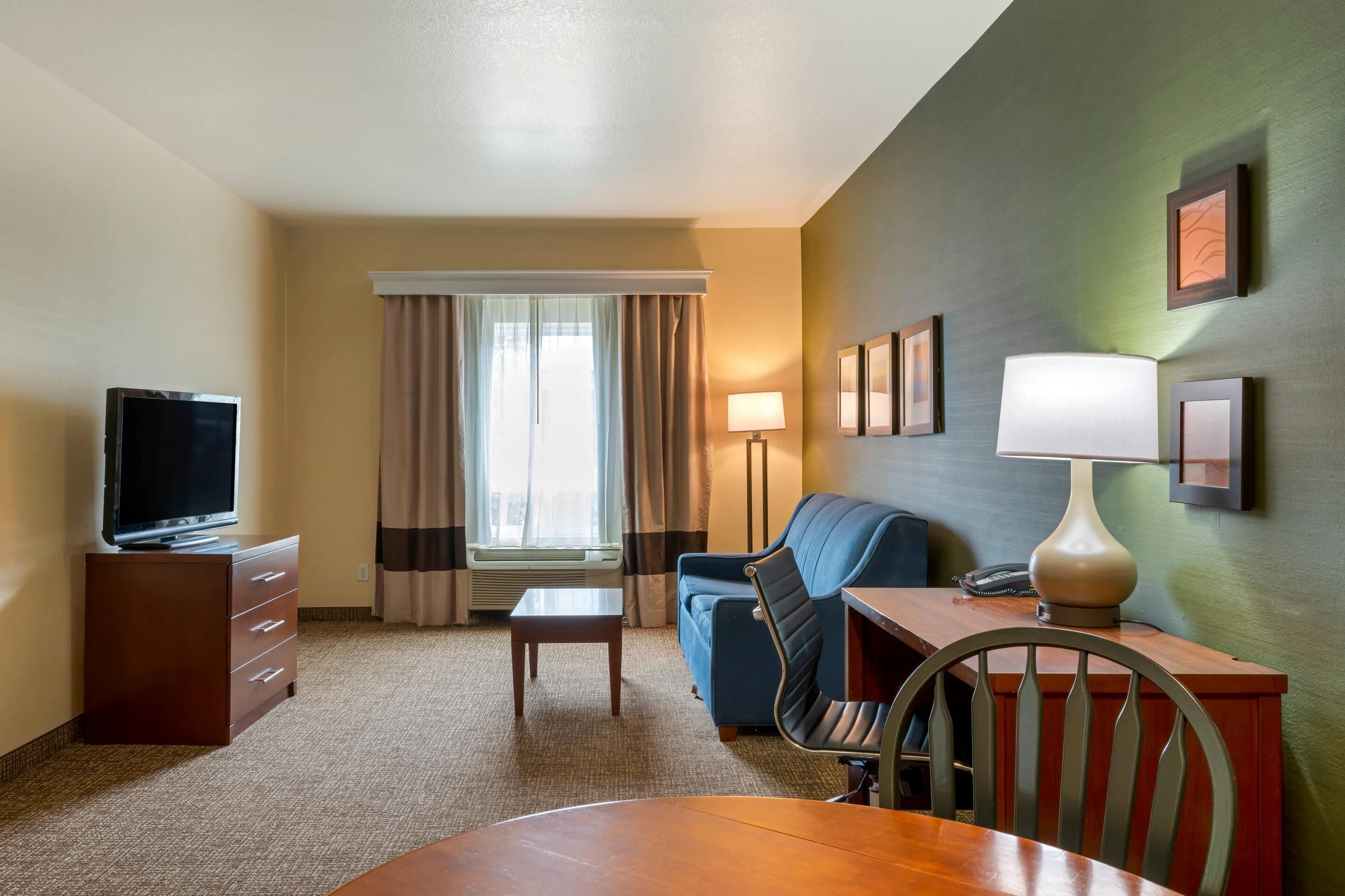 Quarto de hotel com televisão, secretária, cadeira, sofá, abajur e janela com cortinas.