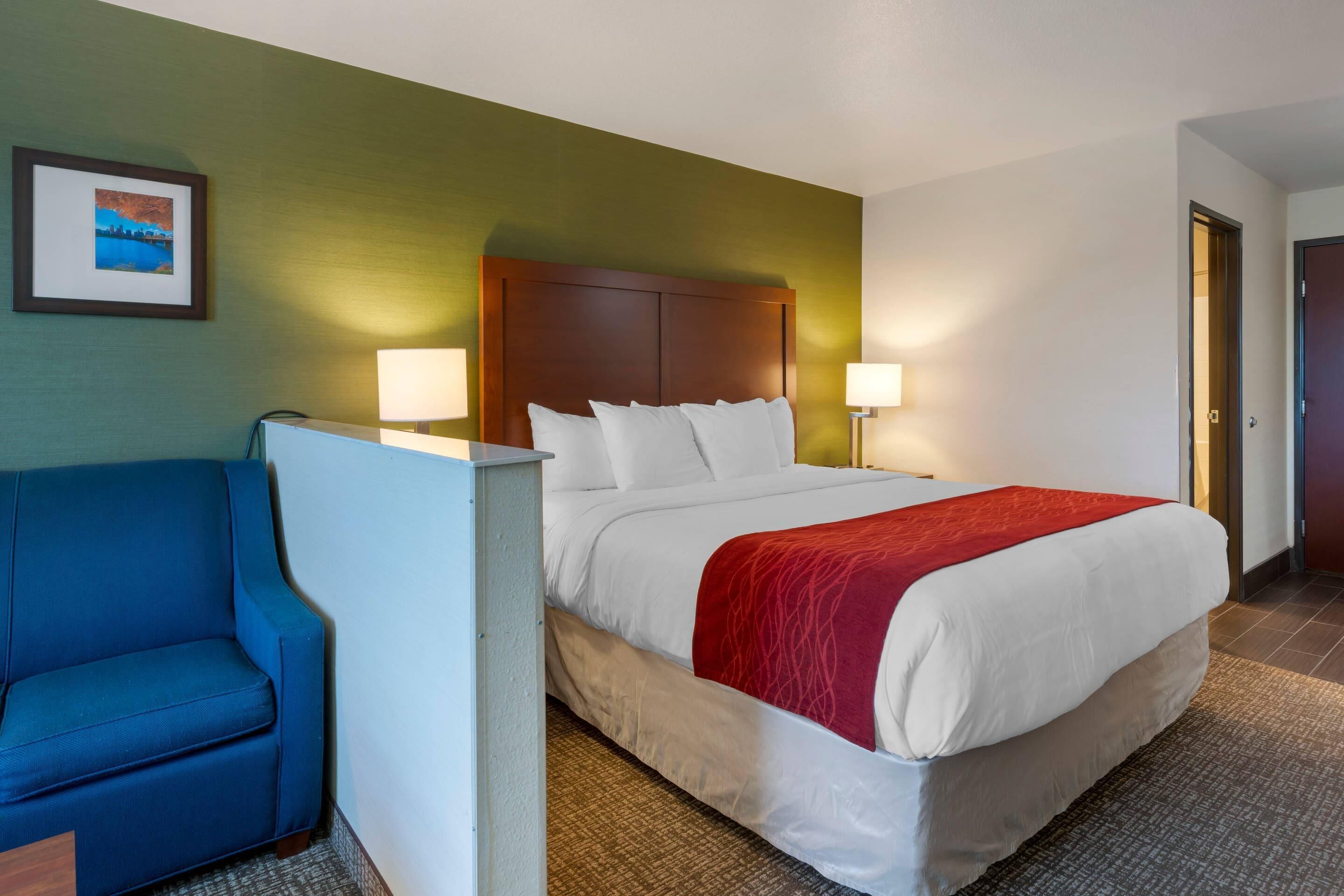 A hotel room with a bed, two bedside lamps, a blue armchair, and a framed picture on the wall.