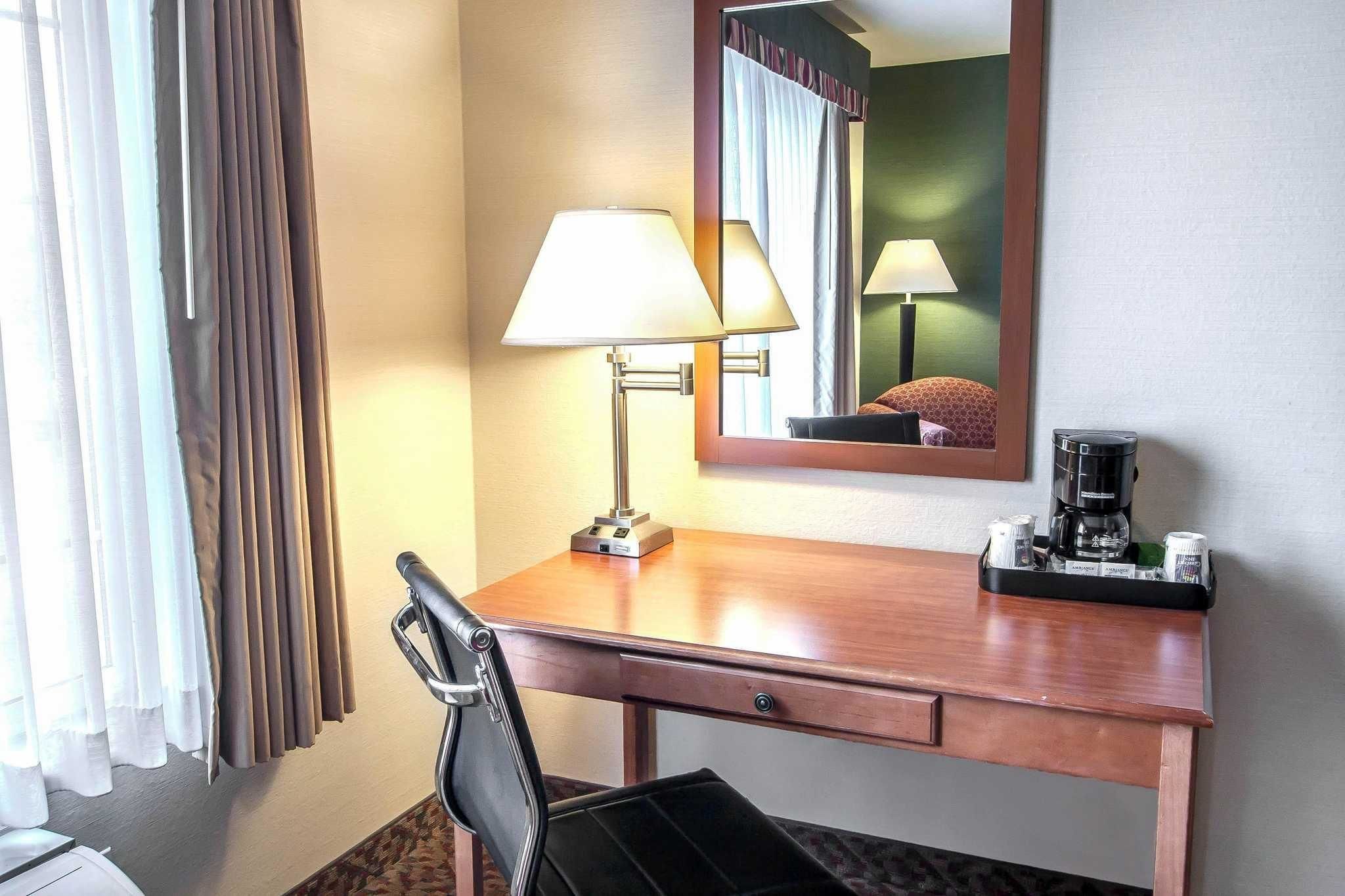 A hotel room with a wooden desk, a chair, a lamp, a coffee maker, and a mirror.