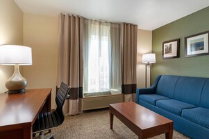 Habitación de hotel con un escritorio de madera, un sofá azul, una silla, una lámpara de pie y una ventana con cortinas.