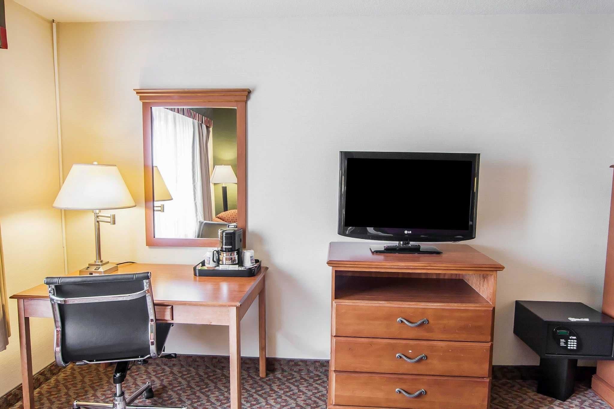 Una camera d'hotel con una scrivania, una sedia, una televisione, uno specchio e un minibar.