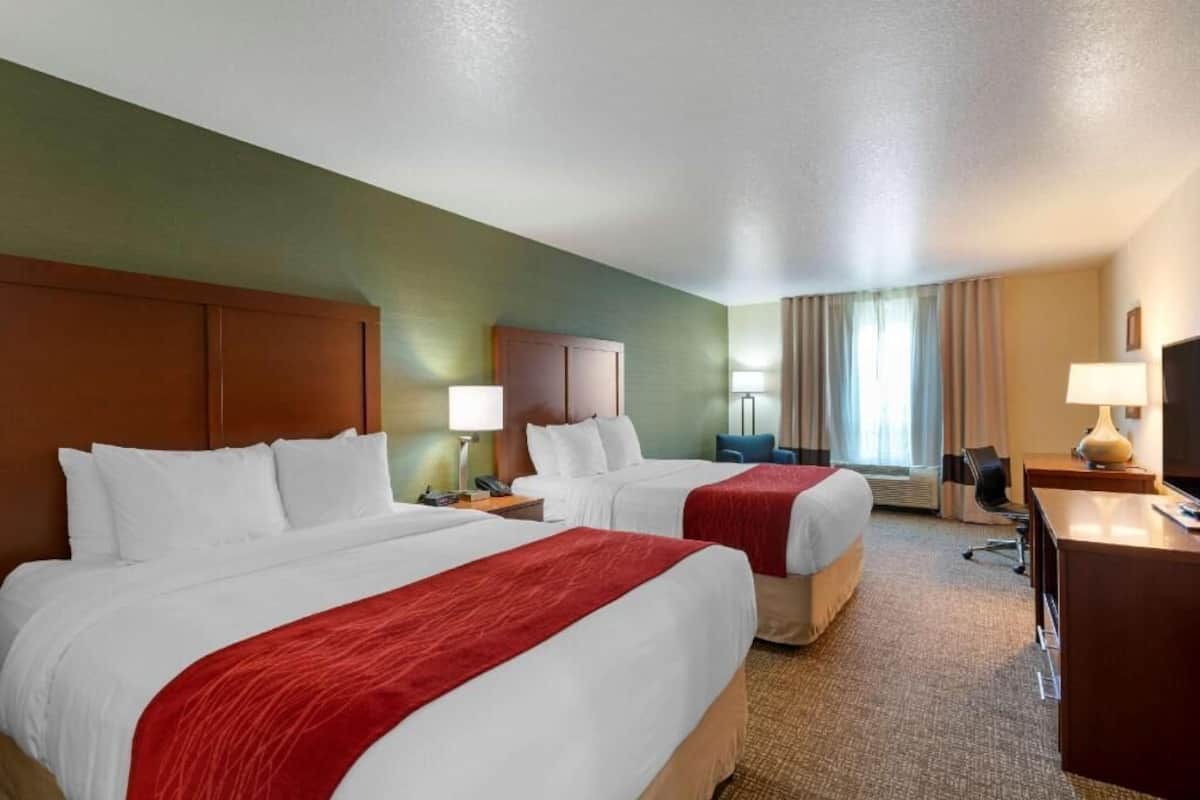Una habitación de hotel con dos camas, un escritorio, una silla, una lámpara y un ventanal con cortinas.