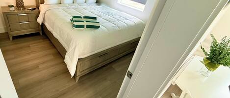 Un dormitorio con una cama de madera, una mesita de noche con una lámpara, una planta pequeña y una ventana con persianas.