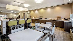 A modern hotel dining area with white tables and chairs, a kitchen in the background, and a high ceiling with recessed lighting. - La Quinta Inn & Suites by Wyndham Manteca - Ripon (Ripon)