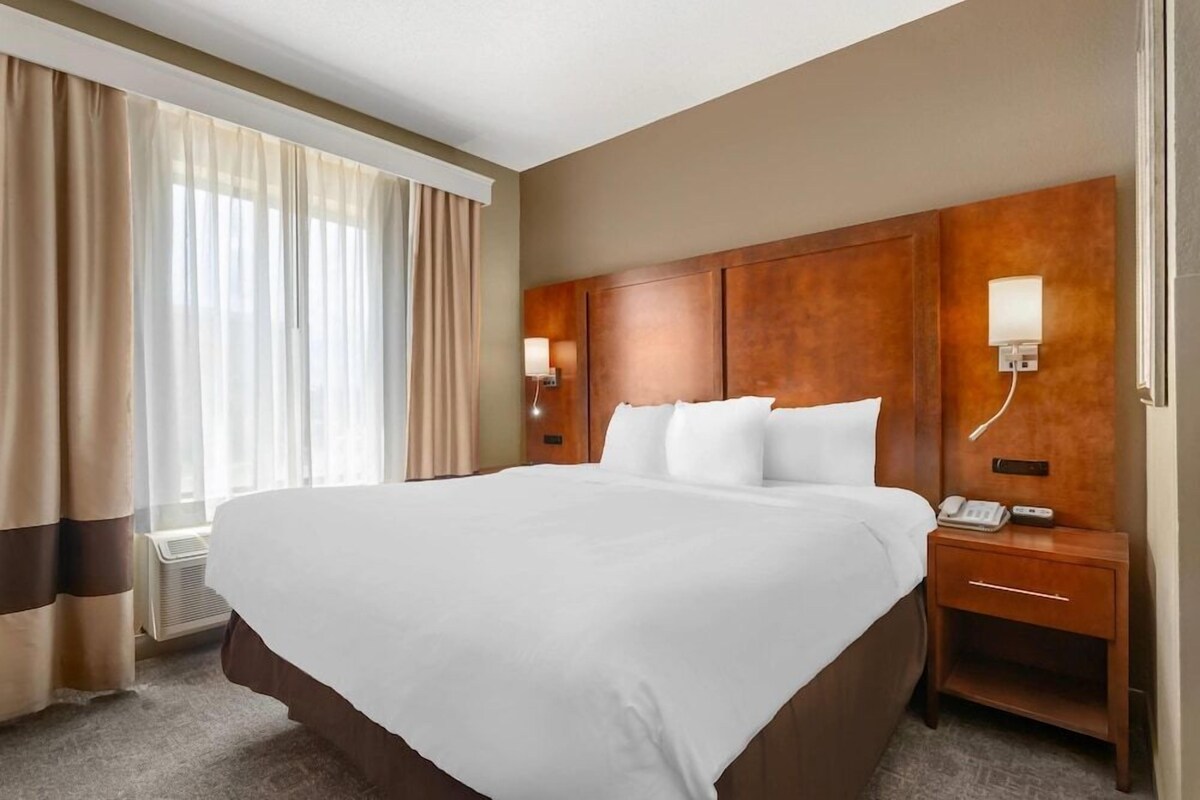 Une chambre d’hôtel avec un grand lit, une tête de lit en bois, une table de chevet avec un téléphone et une fenêtre avec des rideaux.