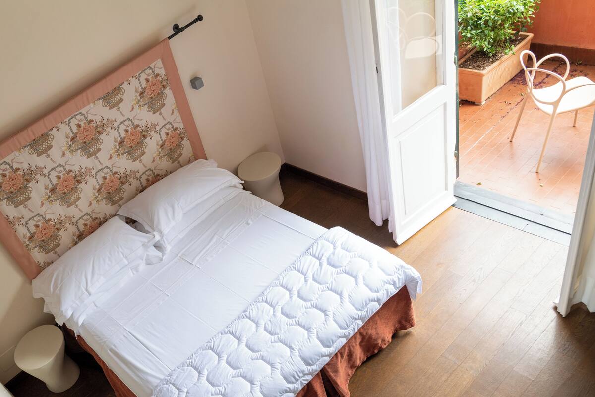 A neatly made bed with a floral headboard, a white bedside table, and a view of a balcony with a potted plant and a chair.
