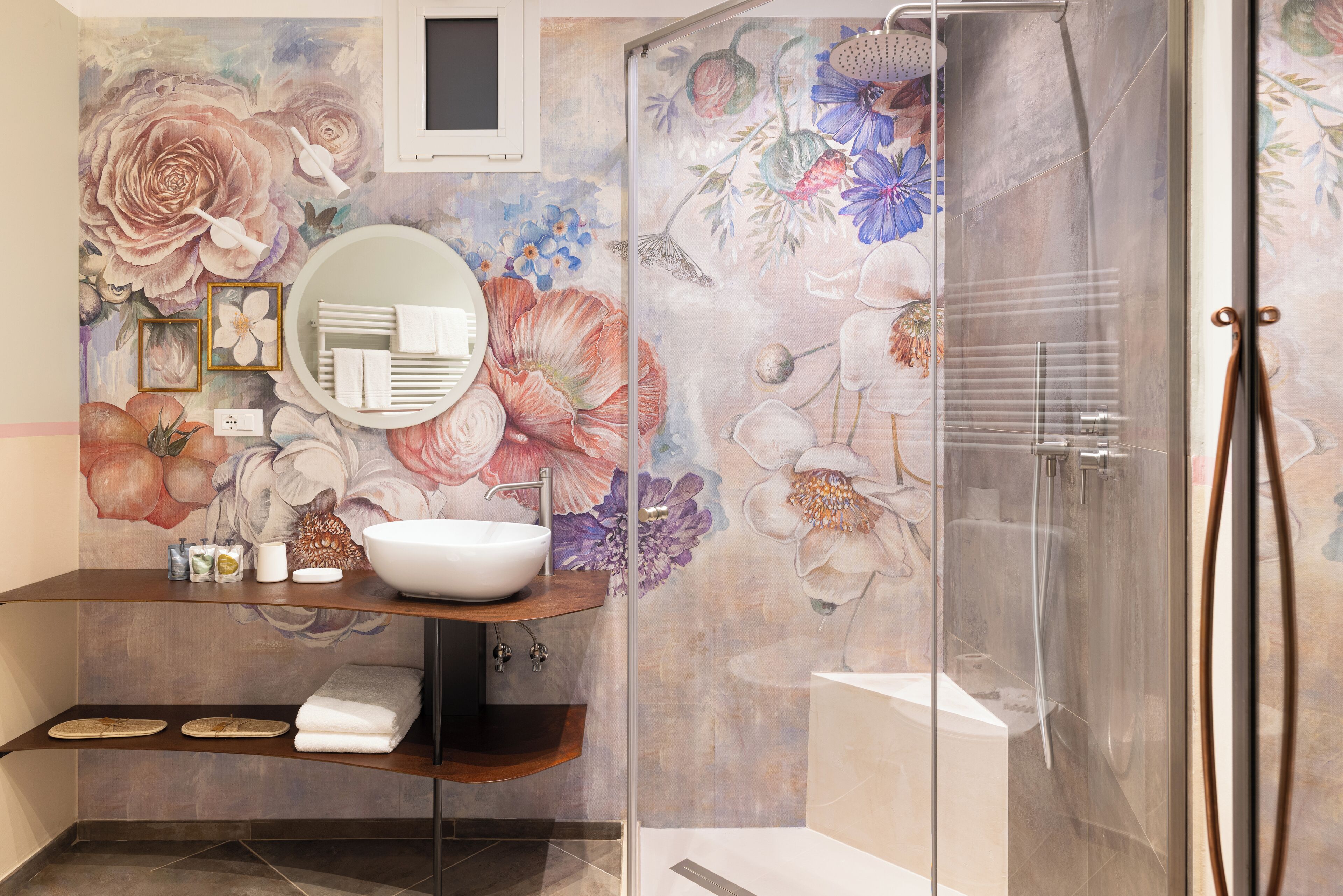 A bathroom with a large floral mural, a round mirror, a sink, and a glass-enclosed shower.