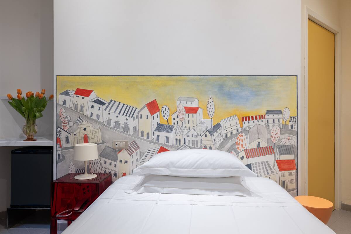A hotel room with a bed, a nightstand with a lamp, a vase of flowers, and a large cityscape mural on the wall.