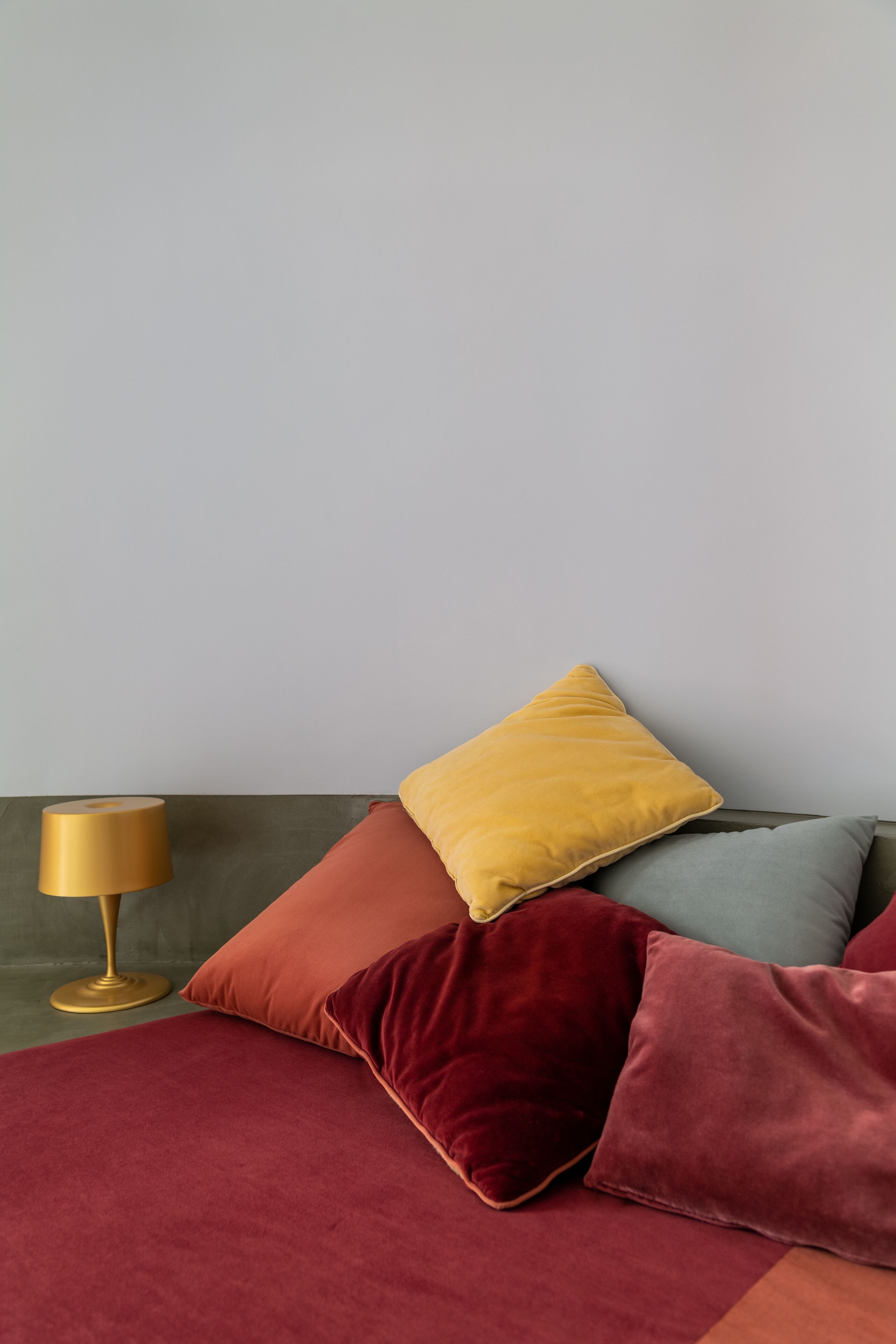 Un lit avec plusieurs oreillers de différentes couleurs, une lampe dorée et un mur sobre.