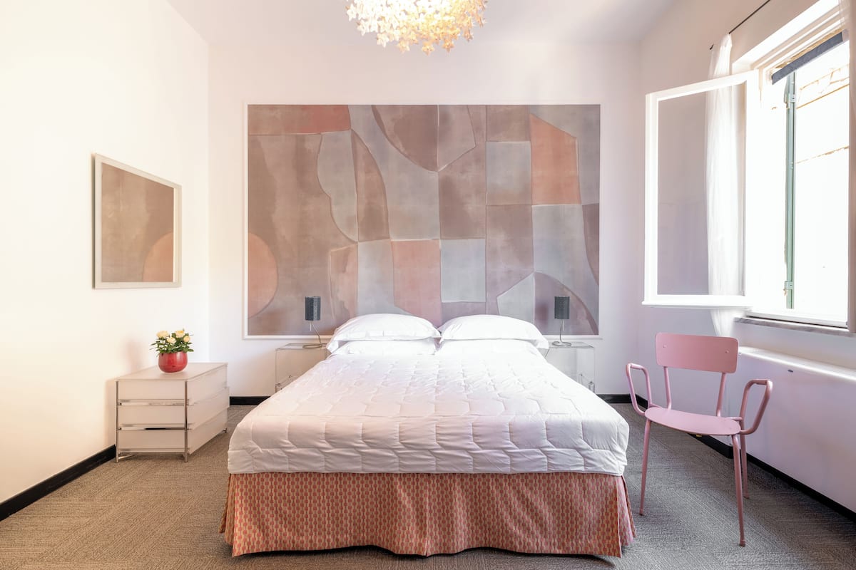 A modern bedroom with a large abstract painting, a bed with a white and pink bedspread, a bedside table with a vase of flowers, and a chair by the window.