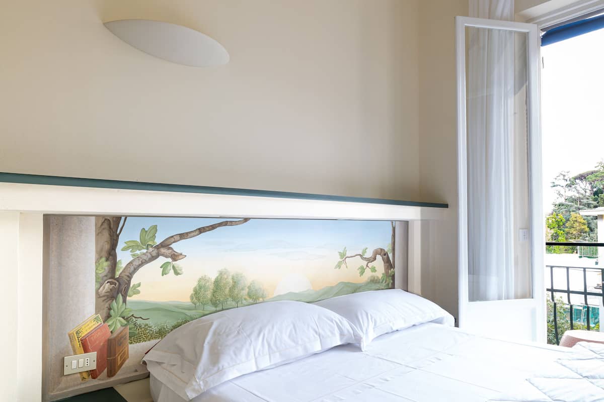 A bedroom with a bed, a nightstand, a painting of a landscape, and a window with curtains.