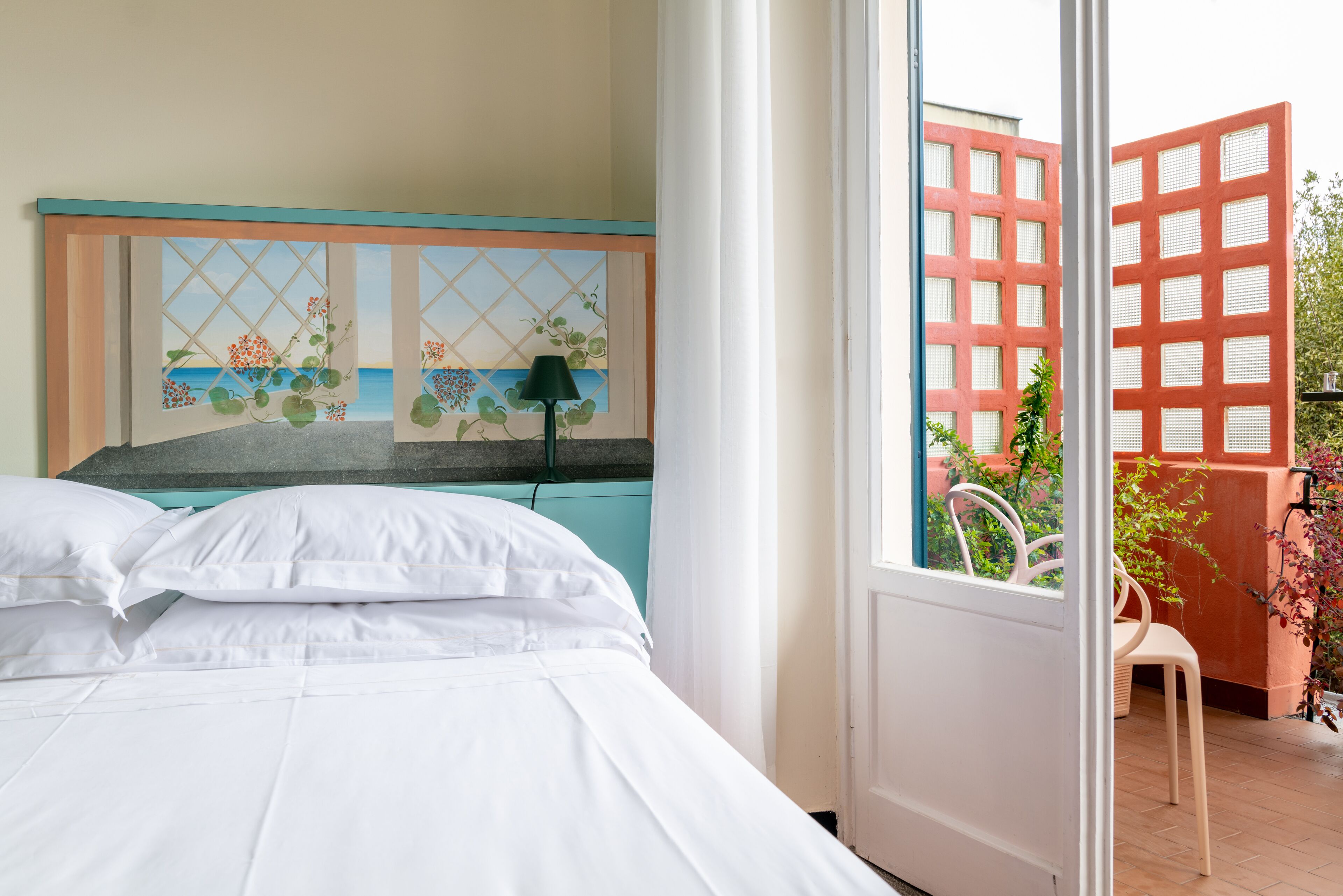 A bedroom with a bed, a window with a view of the sea, and a door leading to an outdoor area with a chair and plants.