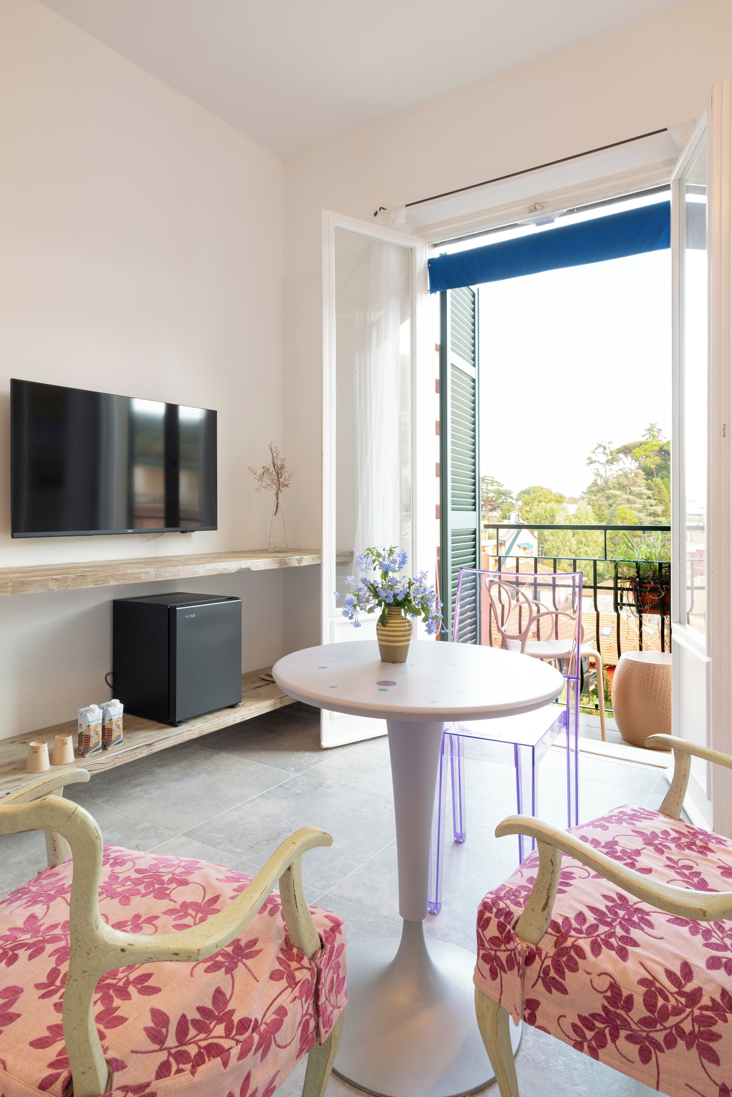Una camera moderna con TV a schermo piatto, un piccolo frigorifero, un tavolo rotondo con sedie e un balcone con vista.