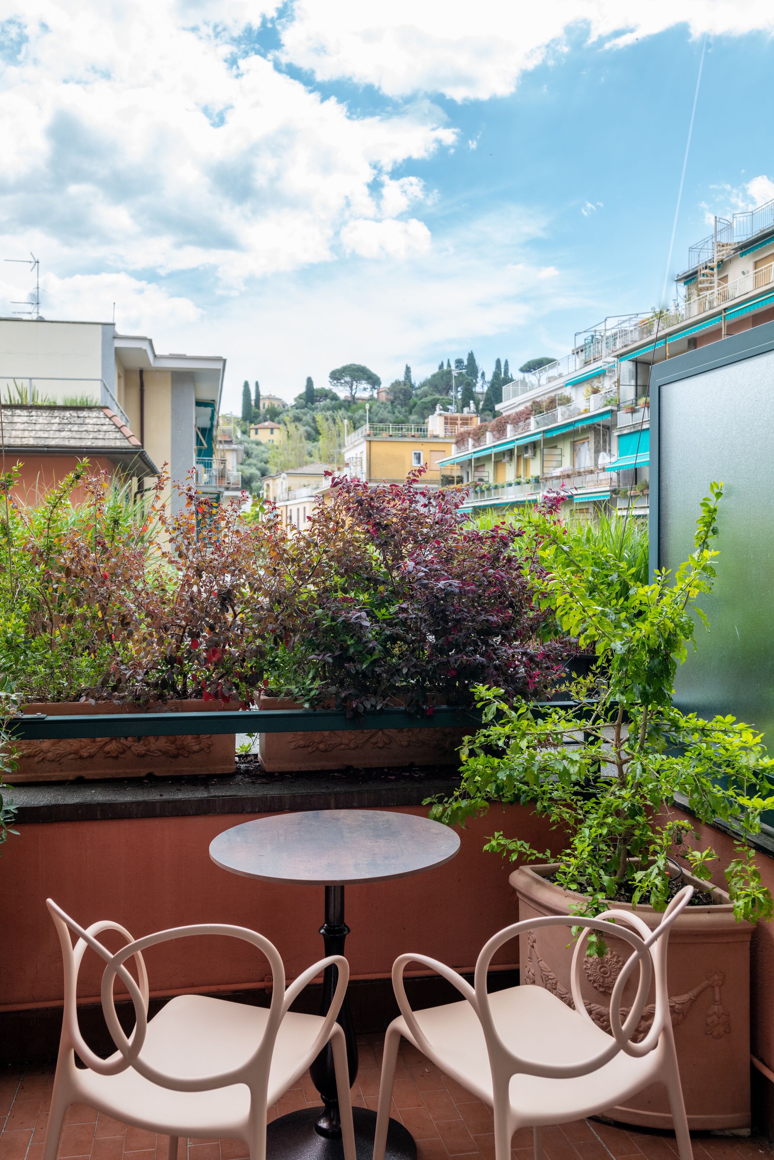 A terrace with two chairs and a table, surrounded by potted plants and overlooking a cityscape.