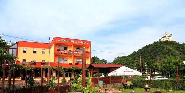 A hotel with a cafe and restaurant, surrounded by greenery and a hill with a structure on top. - Hotel Bellavista (Braubach)