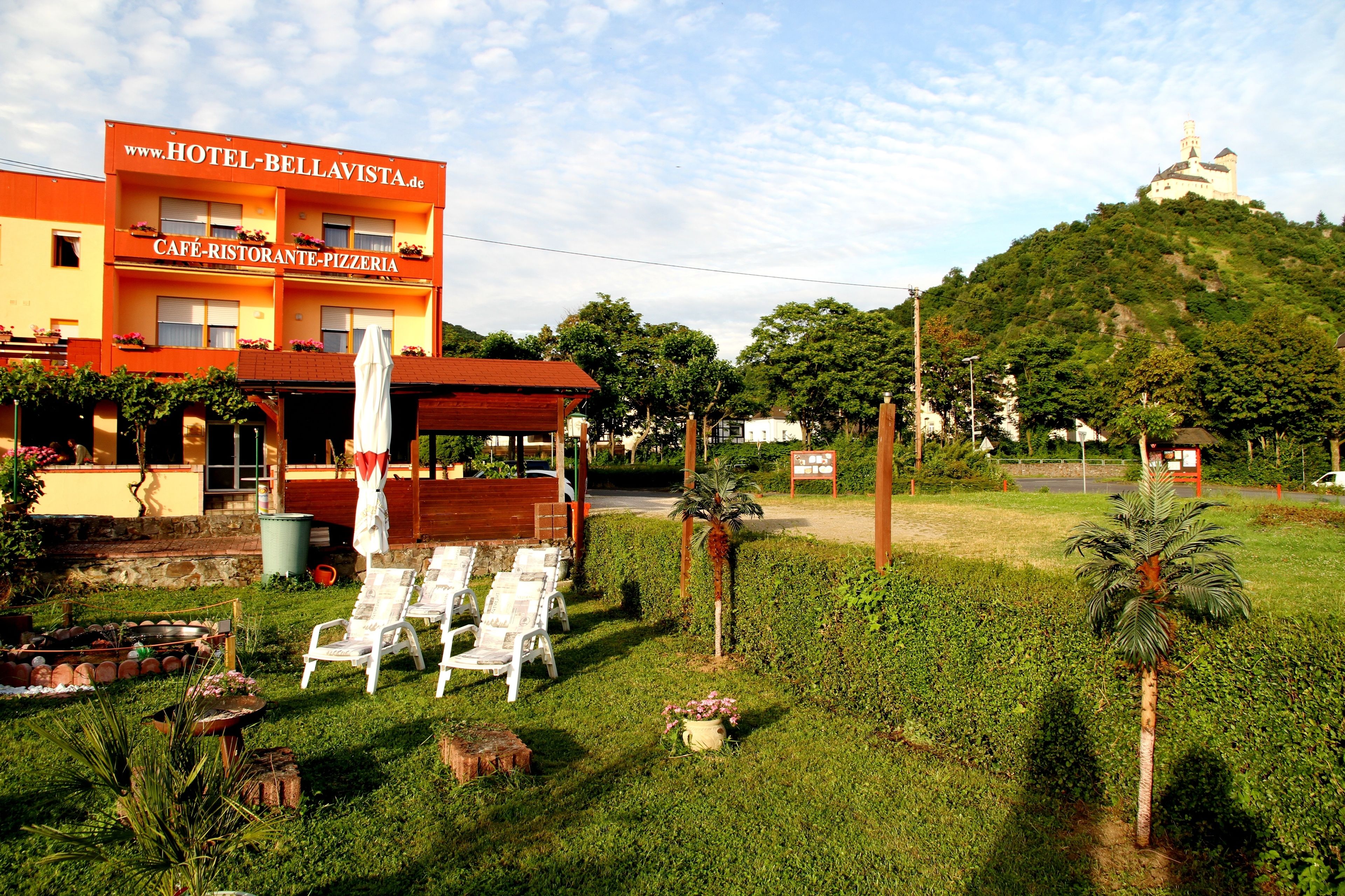 A hotel with a cafe and pizzeria, outdoor seating, and a hill with a castle-like structure in the background.