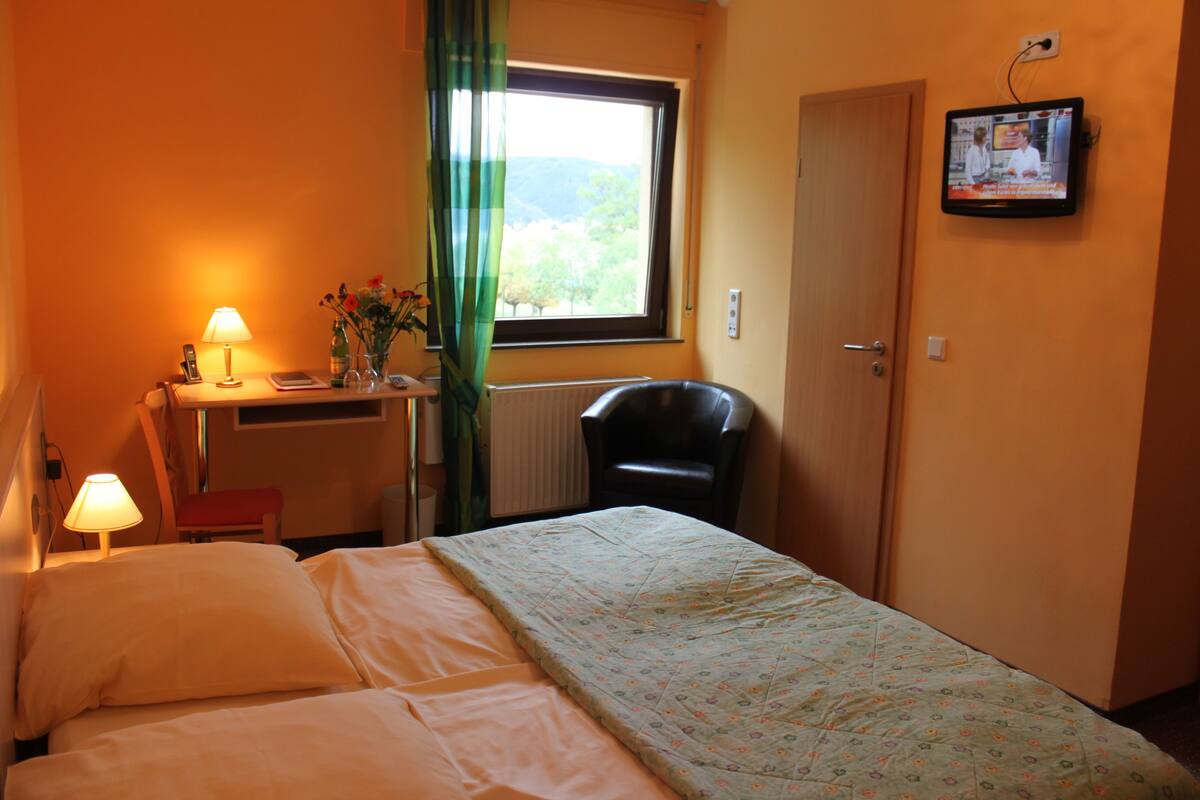 A hotel room with a bed, a desk with a lamp, a chair, a TV on the wall, and a window with curtains.