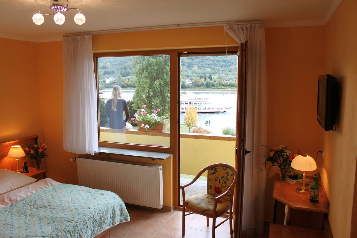 A bedroom with a bed, a chair, a table with a lamp, and a view of a river and trees through a large window.