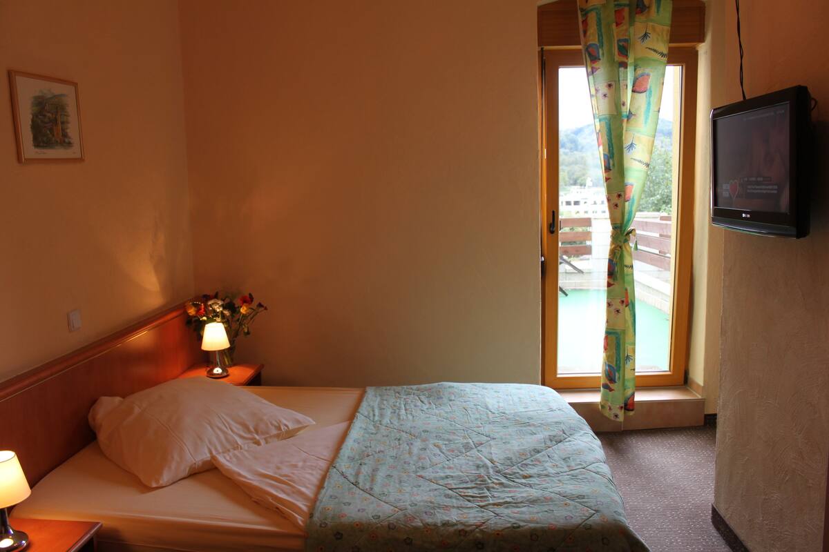 A hotel room with a bed, a TV, a nightstand with a lamp, and a window with a curtain.