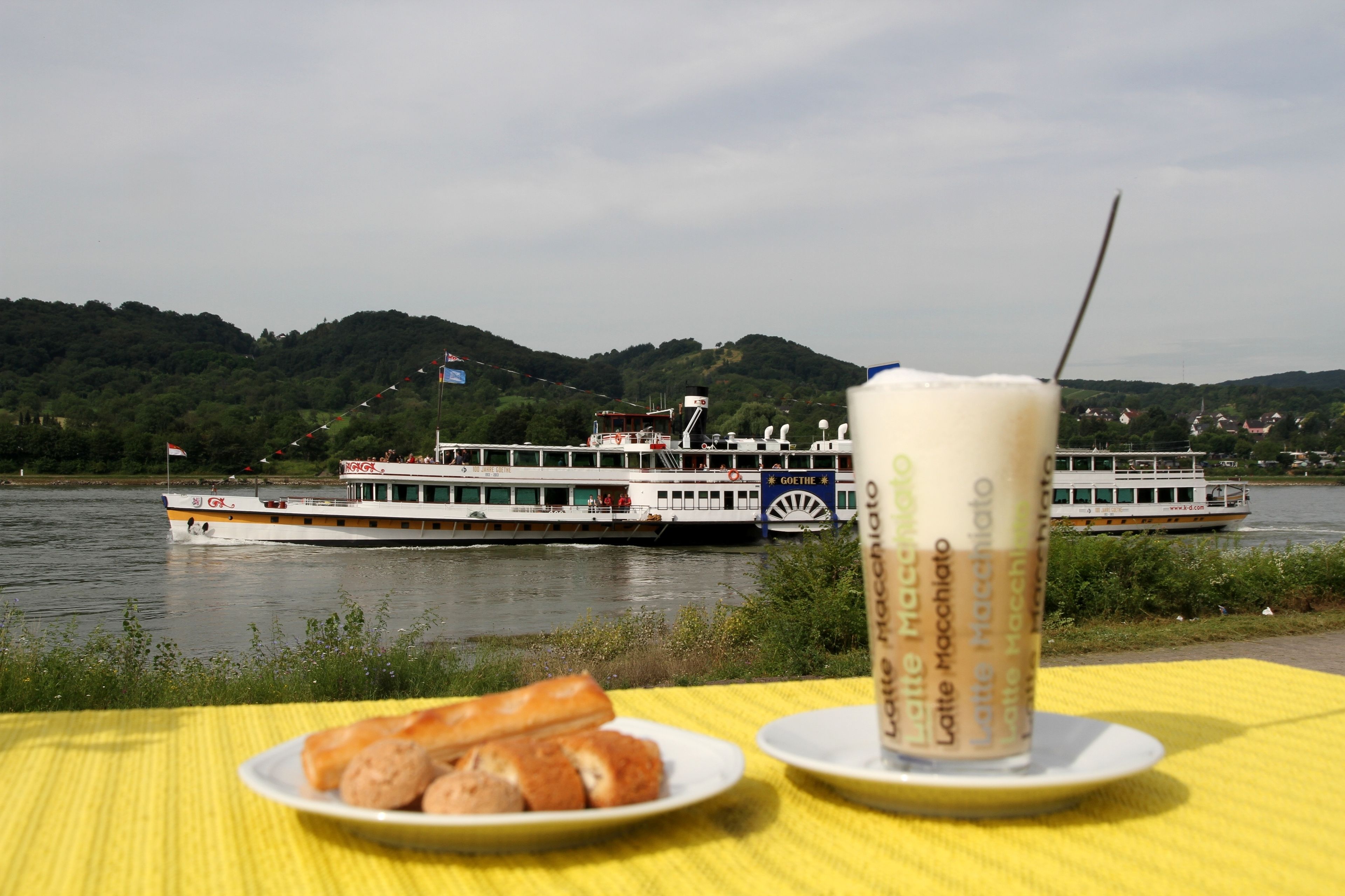 Una mesa con un plato de cruasanes y una taza de latte macchiato, con vistas a un río, una embarcación y colinas al fondo.