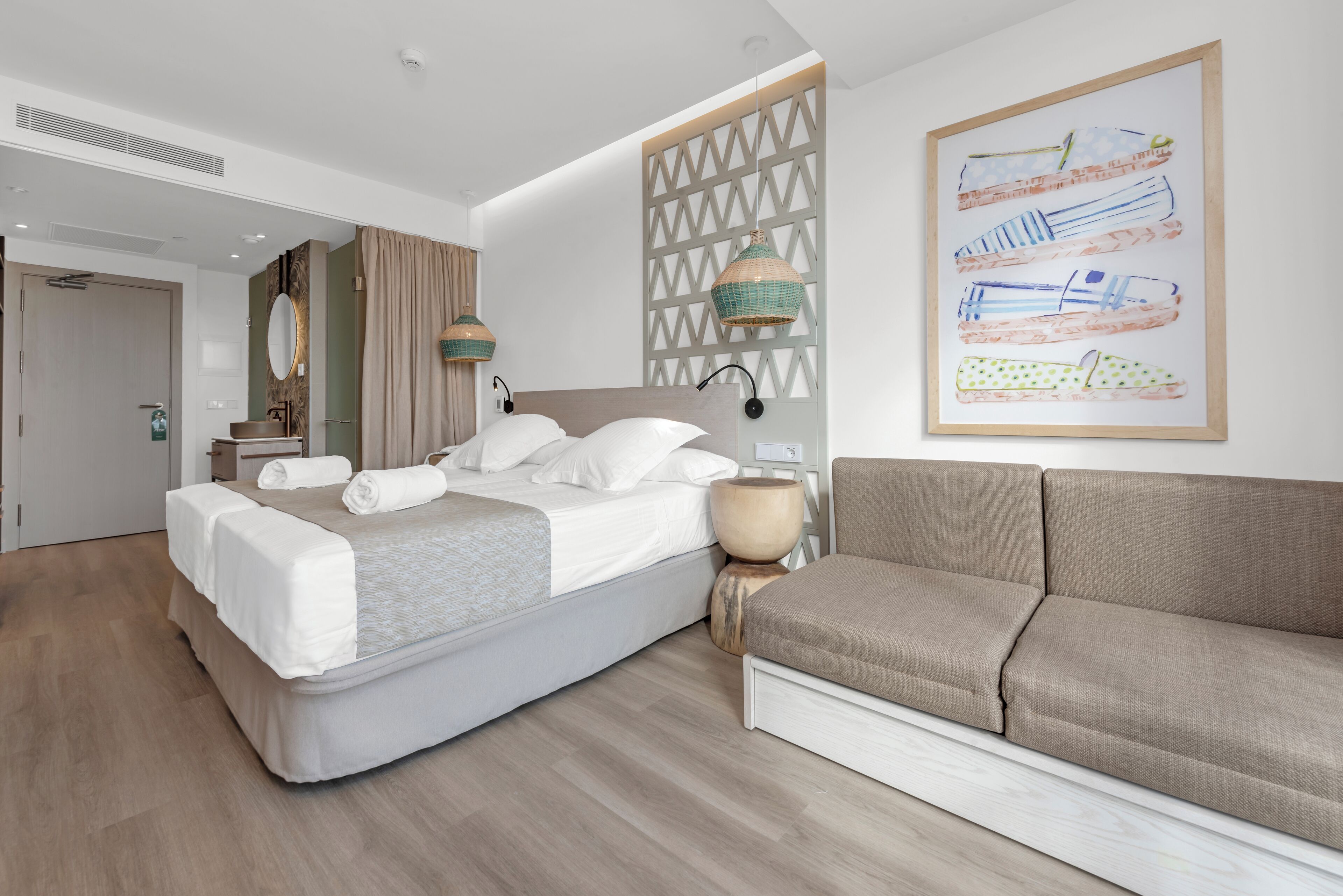 A modern hotel room with a bed, a sofa, a wooden floor, a wall-mounted lamp, and a framed artwork.