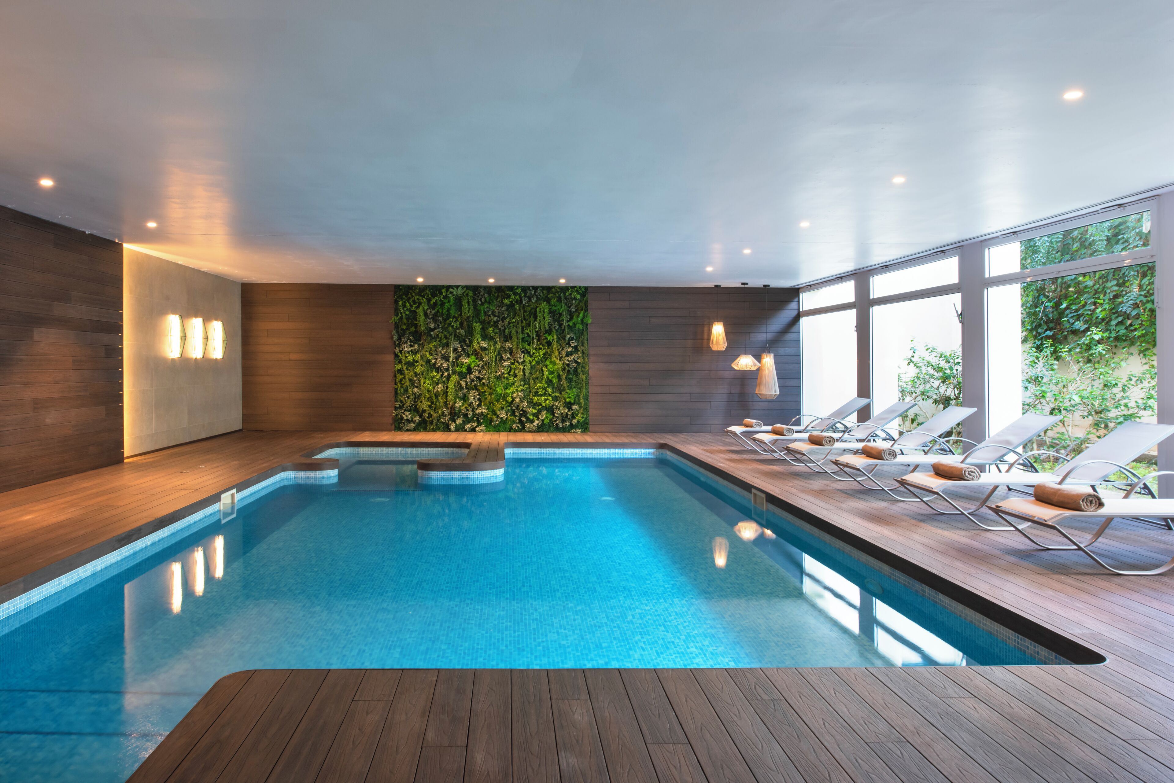 An indoor swimming pool with wooden flooring, a green wall with vertical plants, and sun loungers by the window.