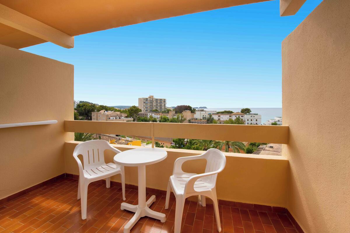 Ein Balkon mit zwei weißen Plastikstühlen und einem kleinen runden Tisch, mit Blick auf eine Stadtlandschaft und das Meer.