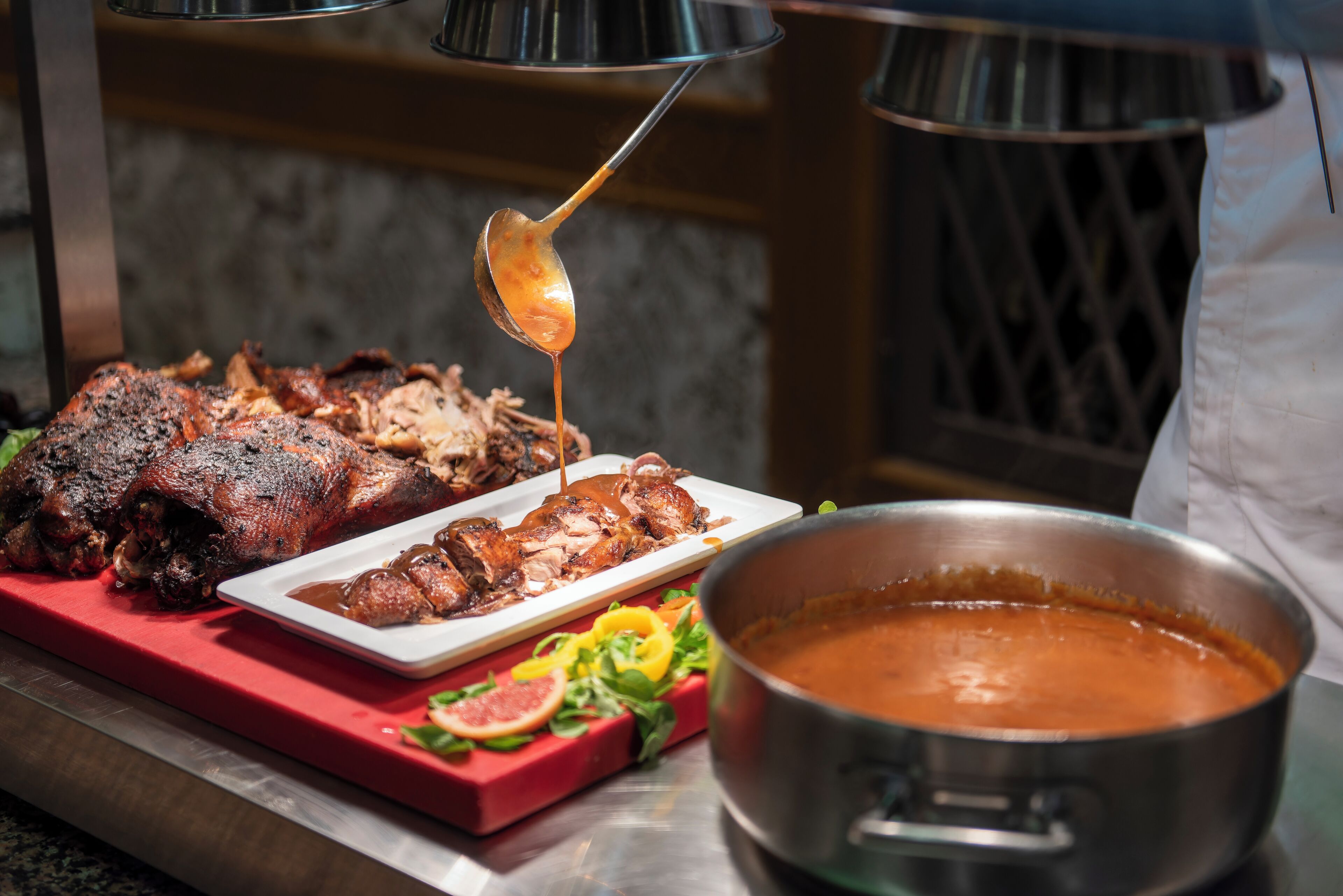Un plat de porc rôti nappé de sauce est servi sur une assiette blanche, avec des tranches de viande à côté, et un pot de sauce est posé sur une planche à découper rouge, entourée de légumes.