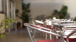 Une rangée de tables à structure métallique, dotées de coussins rouges, est disposée à l’extérieur, agrémentée de plantes en pot, avec un bâtiment en arrière-plan.