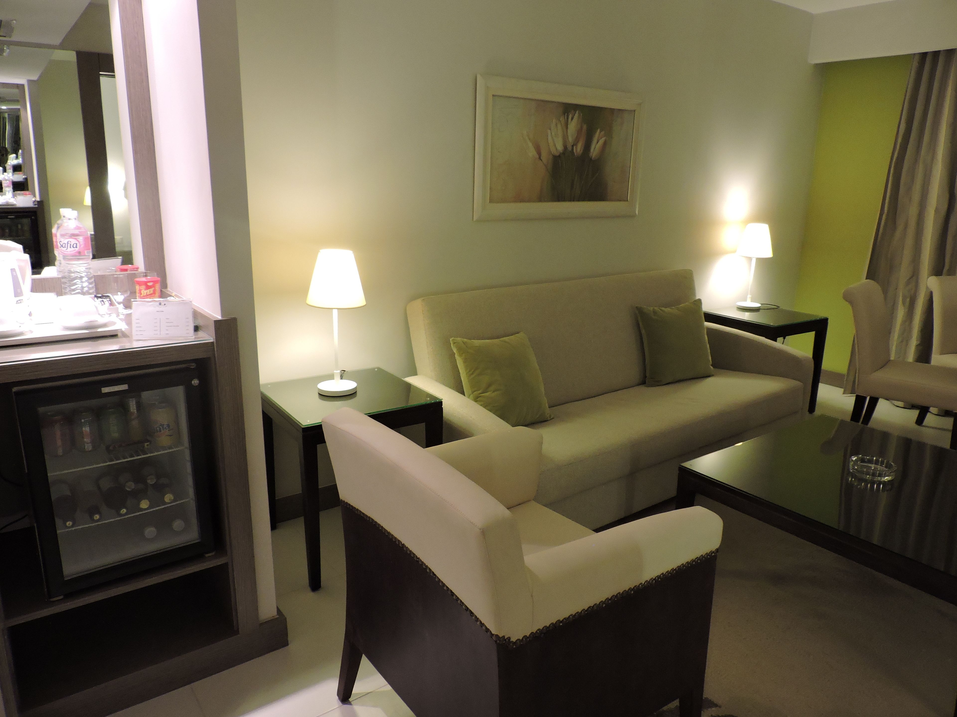 A hotel room with a sofa, two armchairs, a coffee table, a side table with a lamp, a small refrigerator, and a painting on the wall.