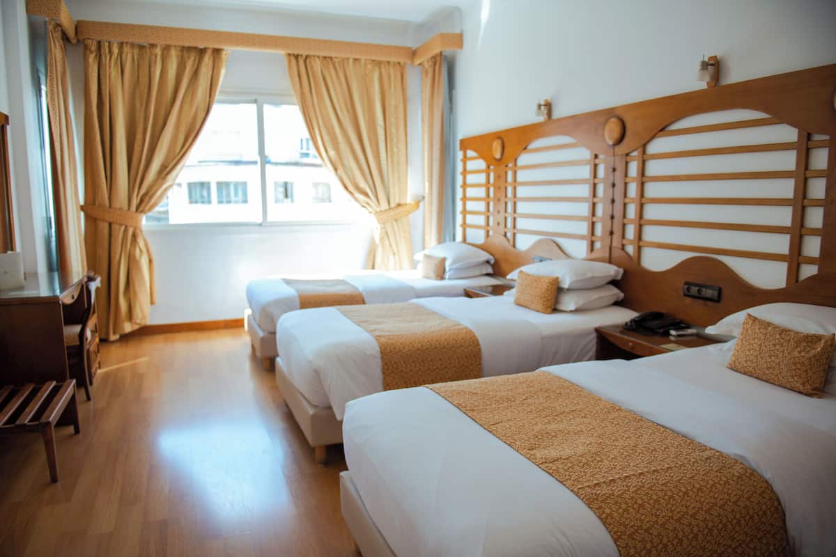Une chambre d’hôtel avec trois lits, des têtes de lit en bois et des rideaux de couleur beige.