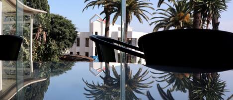 A modern hotel with a glass infinity pool reflecting palm trees and a building.