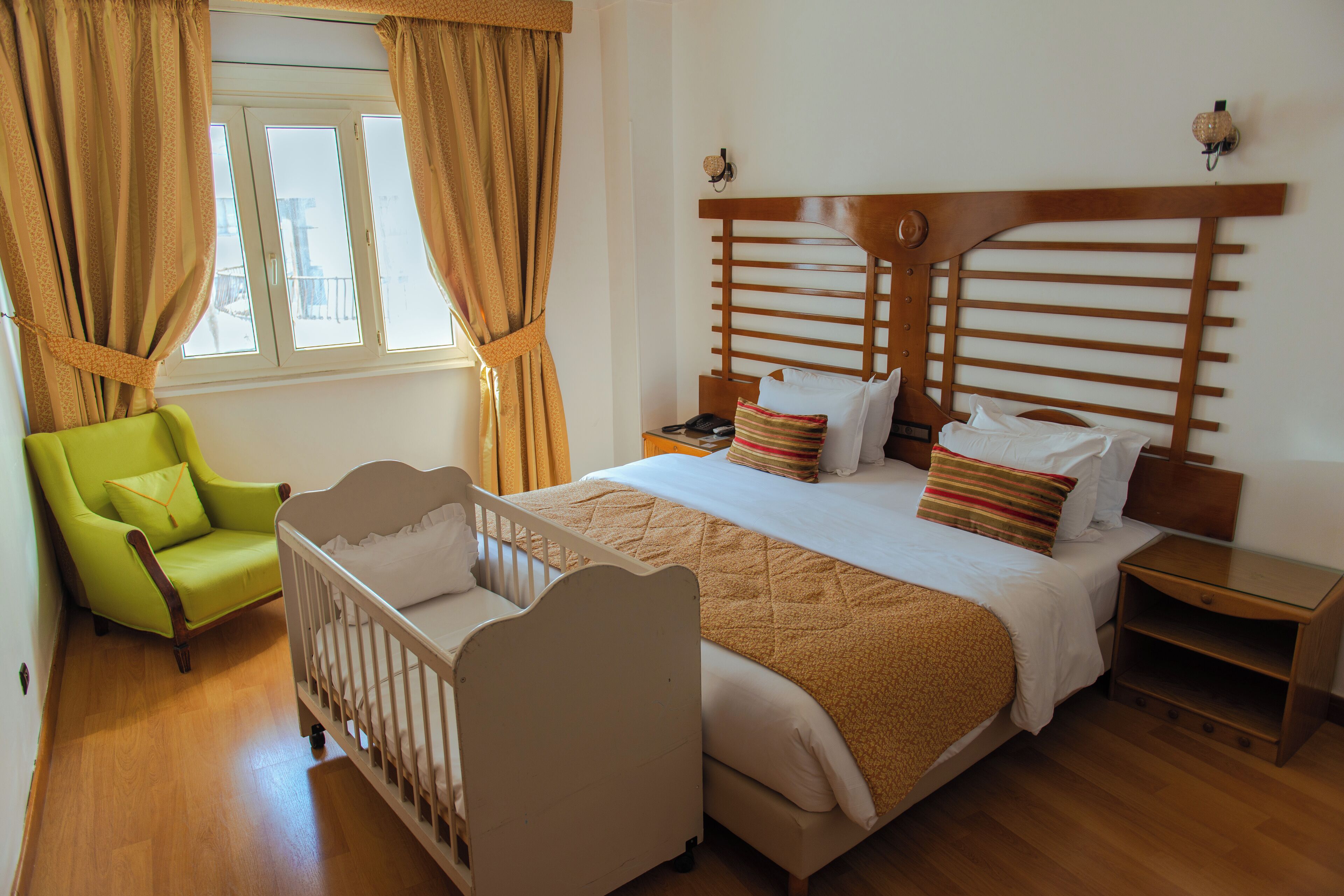 A bedroom with a crib, a green armchair, a wooden headboard, and a bedside table.