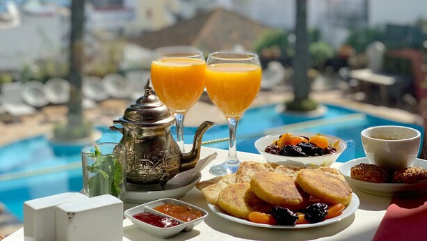 A table with breakfast items, including toast, fruit, and orange juice, overlooking a pool with palm trees and a view of the sea. - Hotel Rembrandt (Tangier)