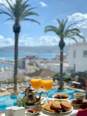 Una mesa con alimentos para el desayuno, incluyendo tostadas, fruta y jugo de naranja, con vista a una piscina con palmeras y al mar.