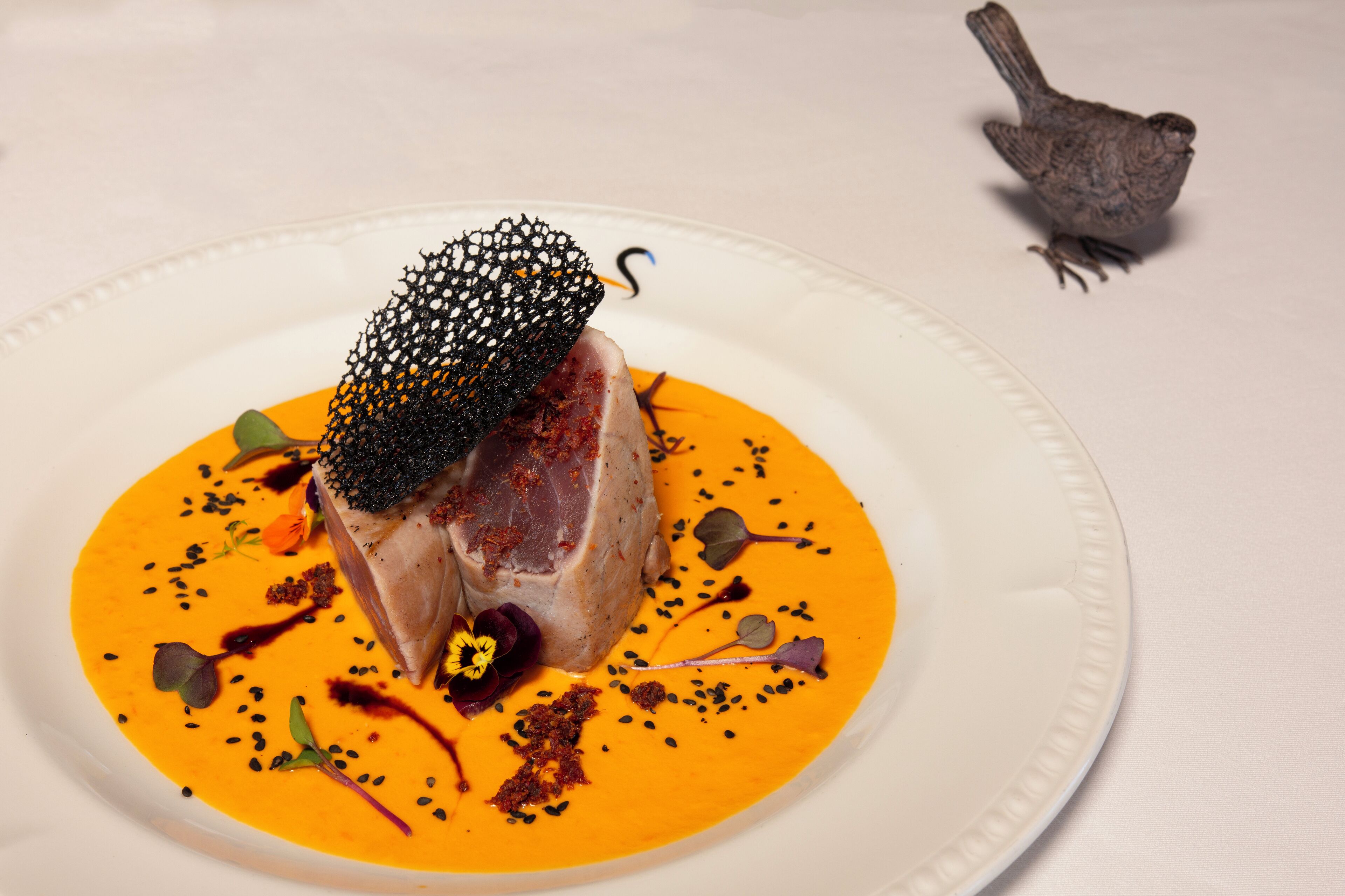 Un plato blanco con carne, flores comestibles negras y salsa anaranjada.