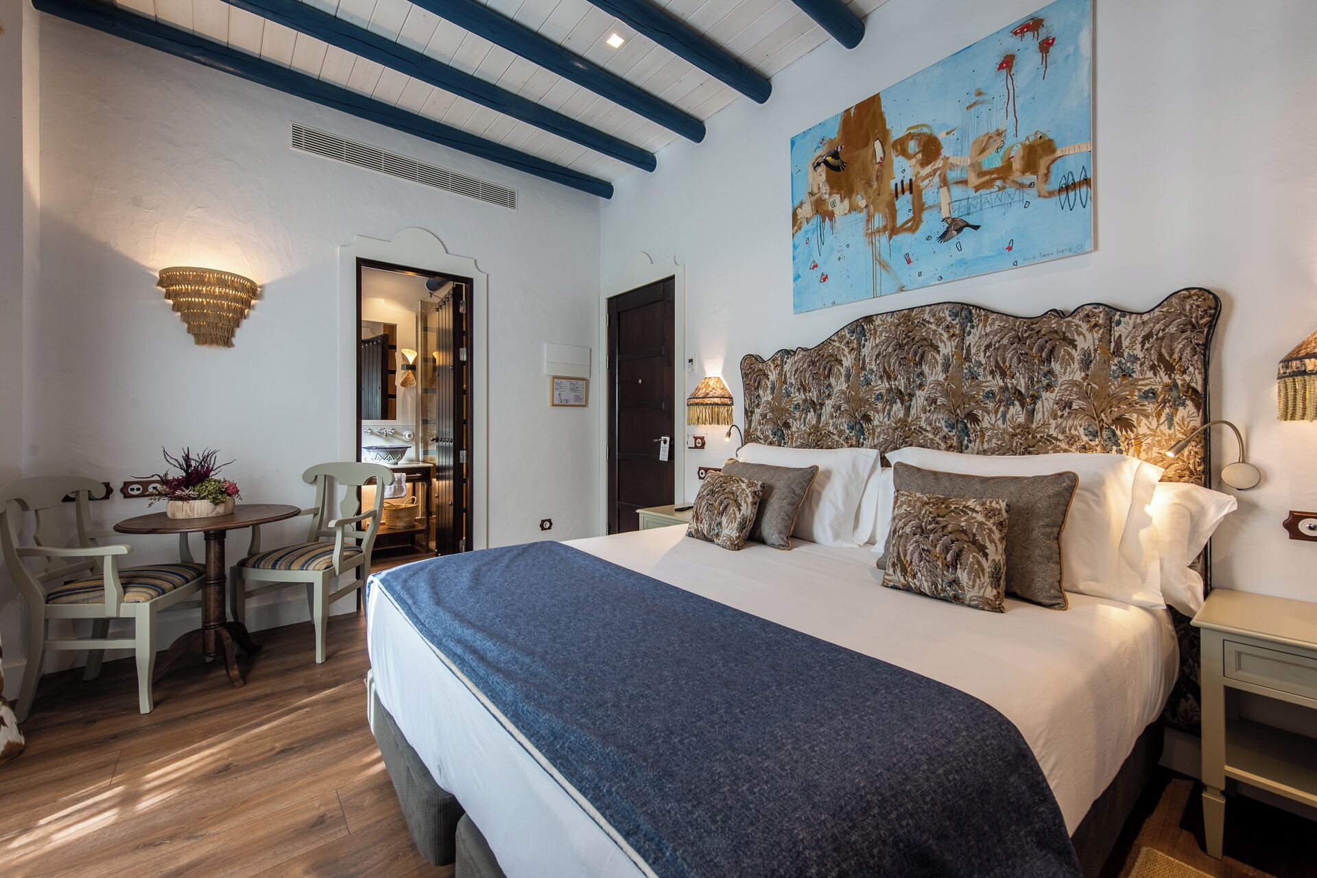 Un dormitorio con una cama grande, una zona de comedor con sillas, una mesa y un cuadro en la pared.
